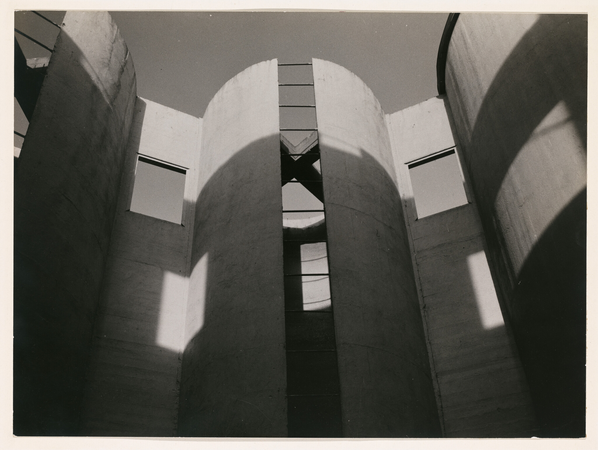 Black and white photograph of a modern architectural structure with curved concrete walls and rectangular windows casting shadows in the sunlight.