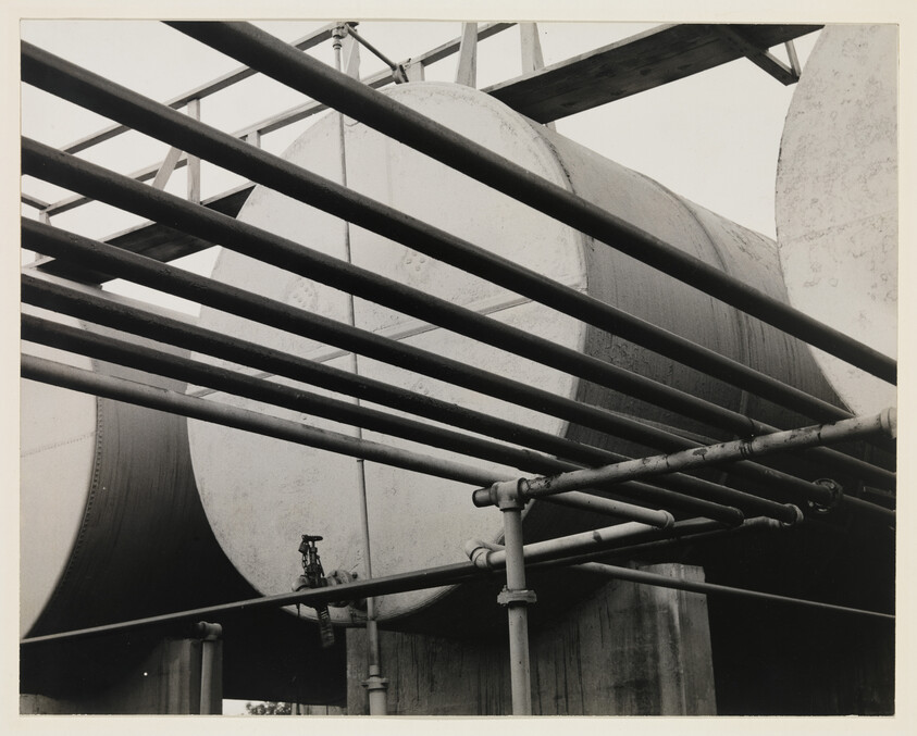 Black and white photo of industrial pipes and scaffolding against large storage tanks.