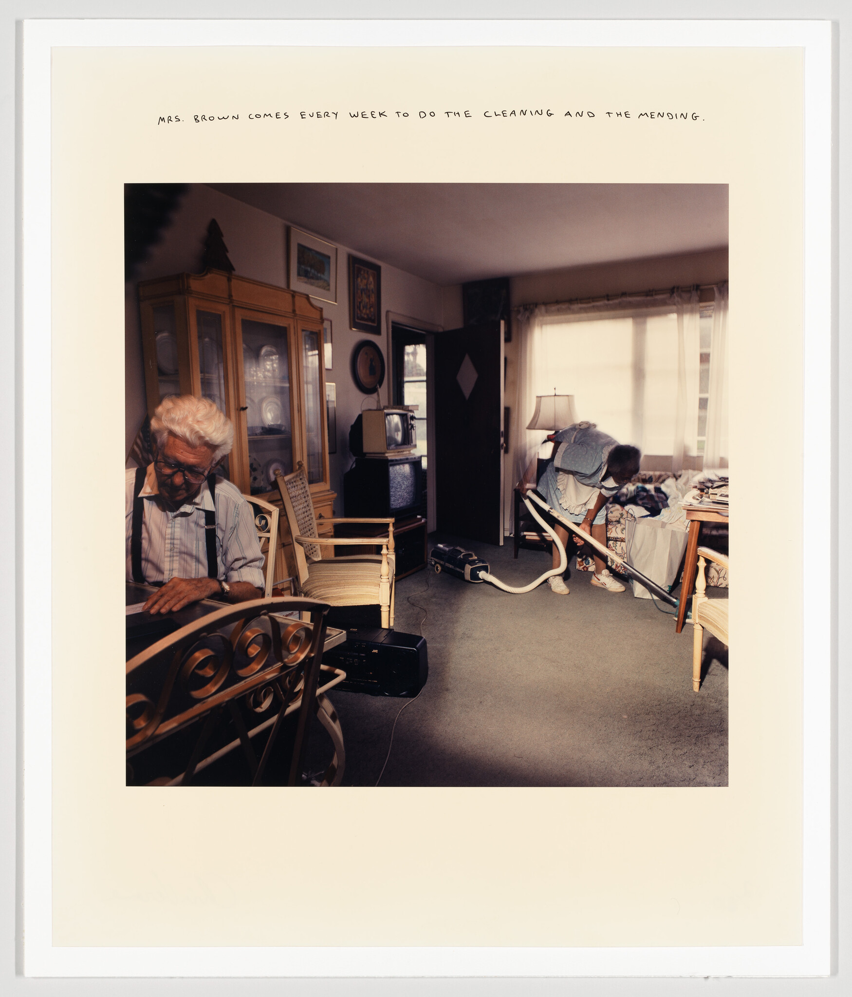 An elderly man sits while a housekeeper vacuums the living room, cleaning and mending weekly.