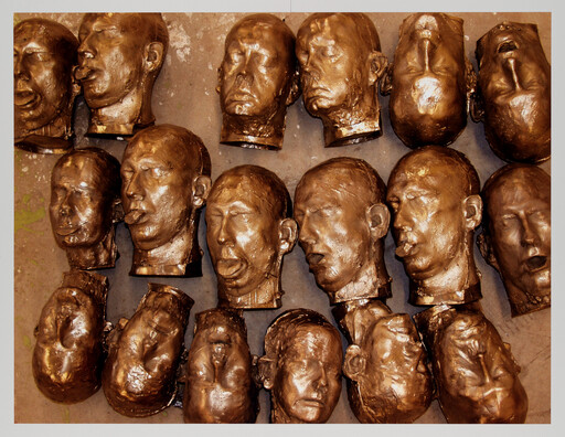 Several bronze-colored sculpted human heads with closed eyes arranged in rows on a surface.