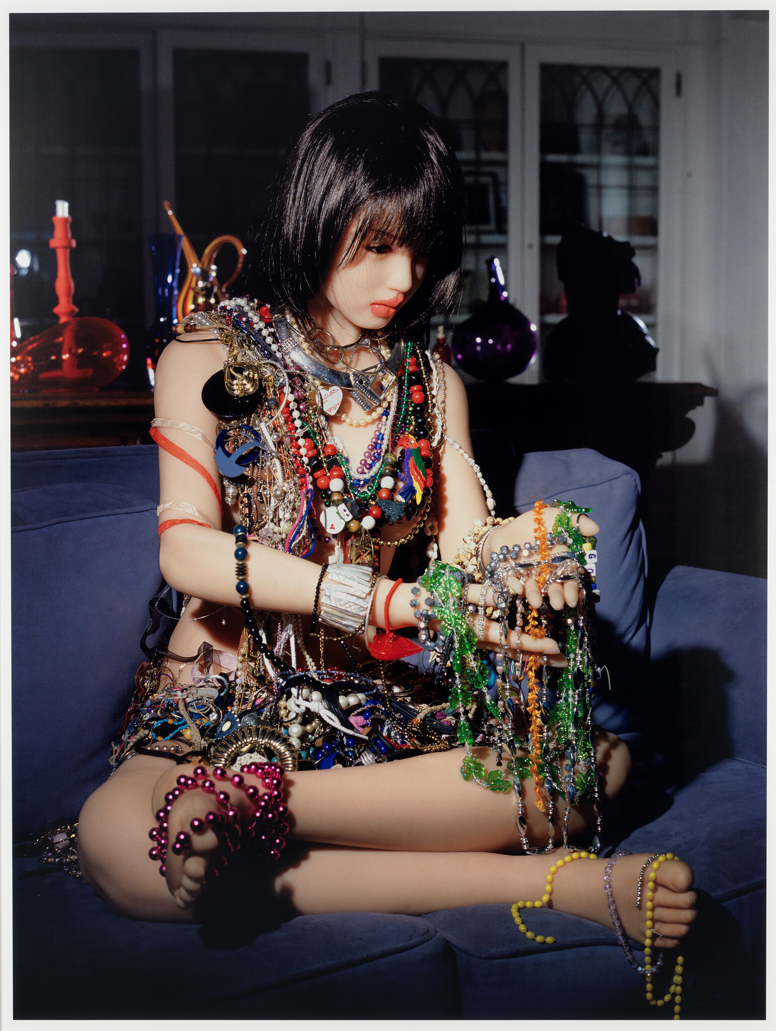 A seated doll with short dark hair wearing and holding layers of colorful necklaces and bracelets.