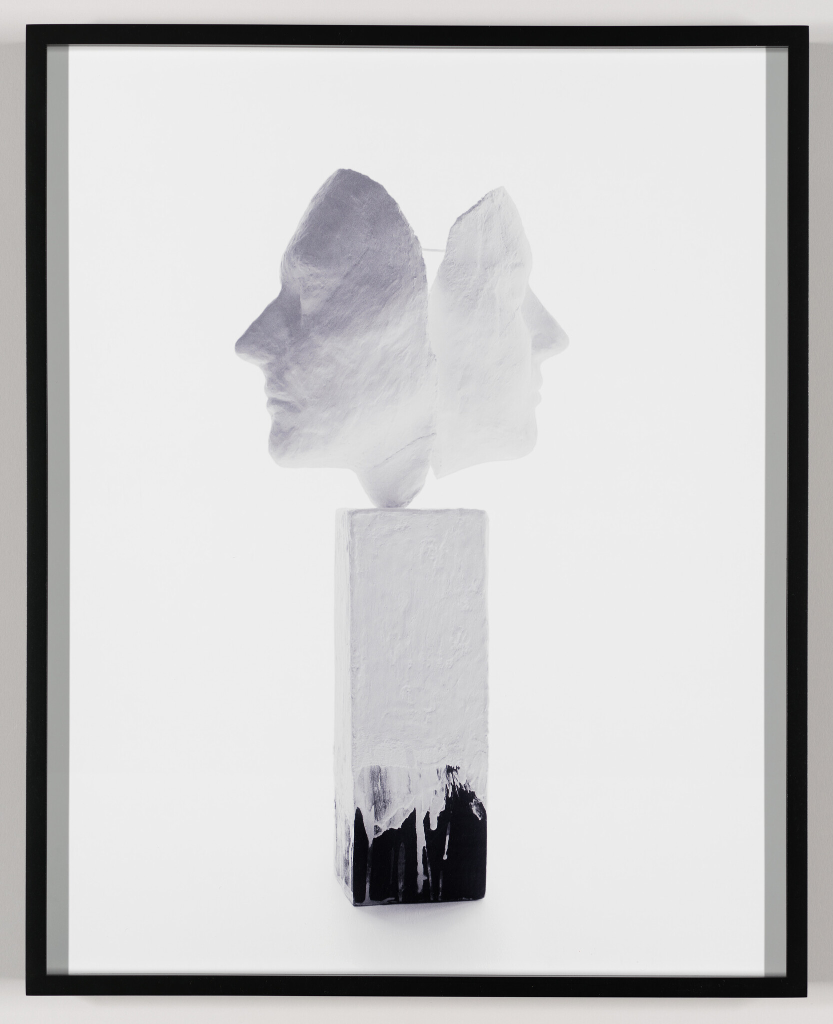 White double-faced sculpture mounted on a tall rectangular pedestal with black paint drips.
