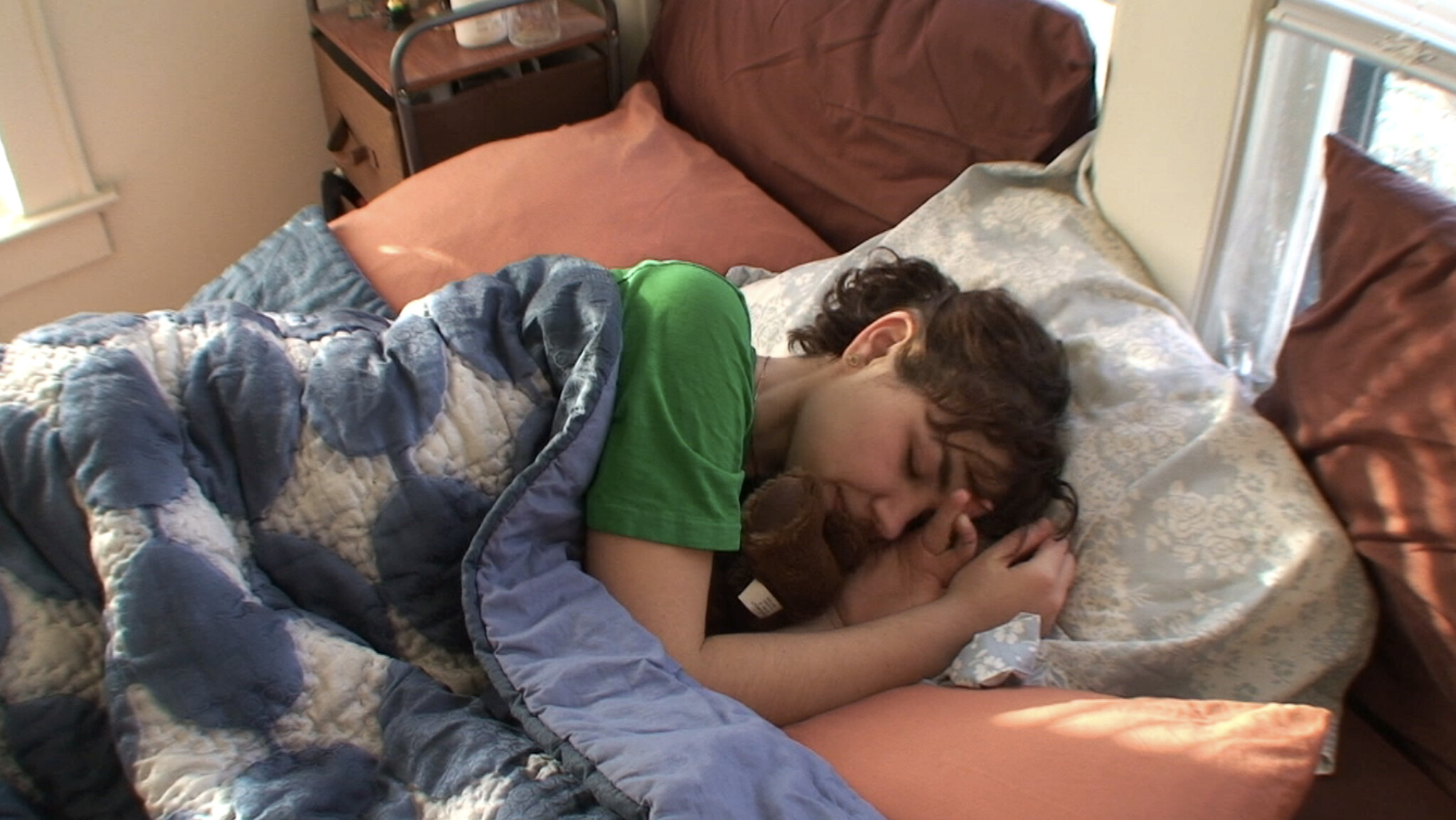 A person sleeps curled on a bed, hugging a small stuffed animal under a blue quilt.