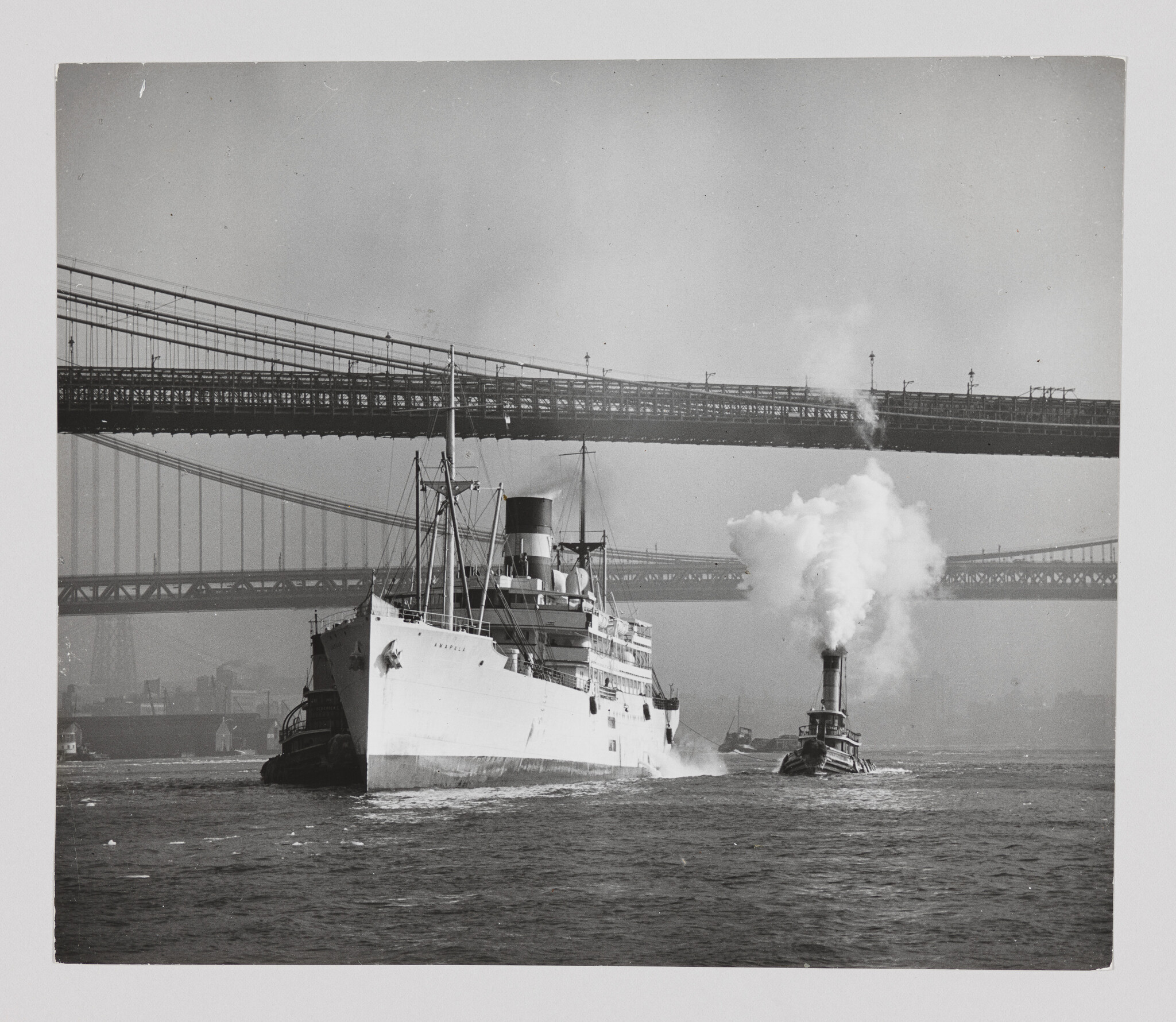 A large passenger ship is being guided by a smoke-emitting tugboat under suspension bridges.