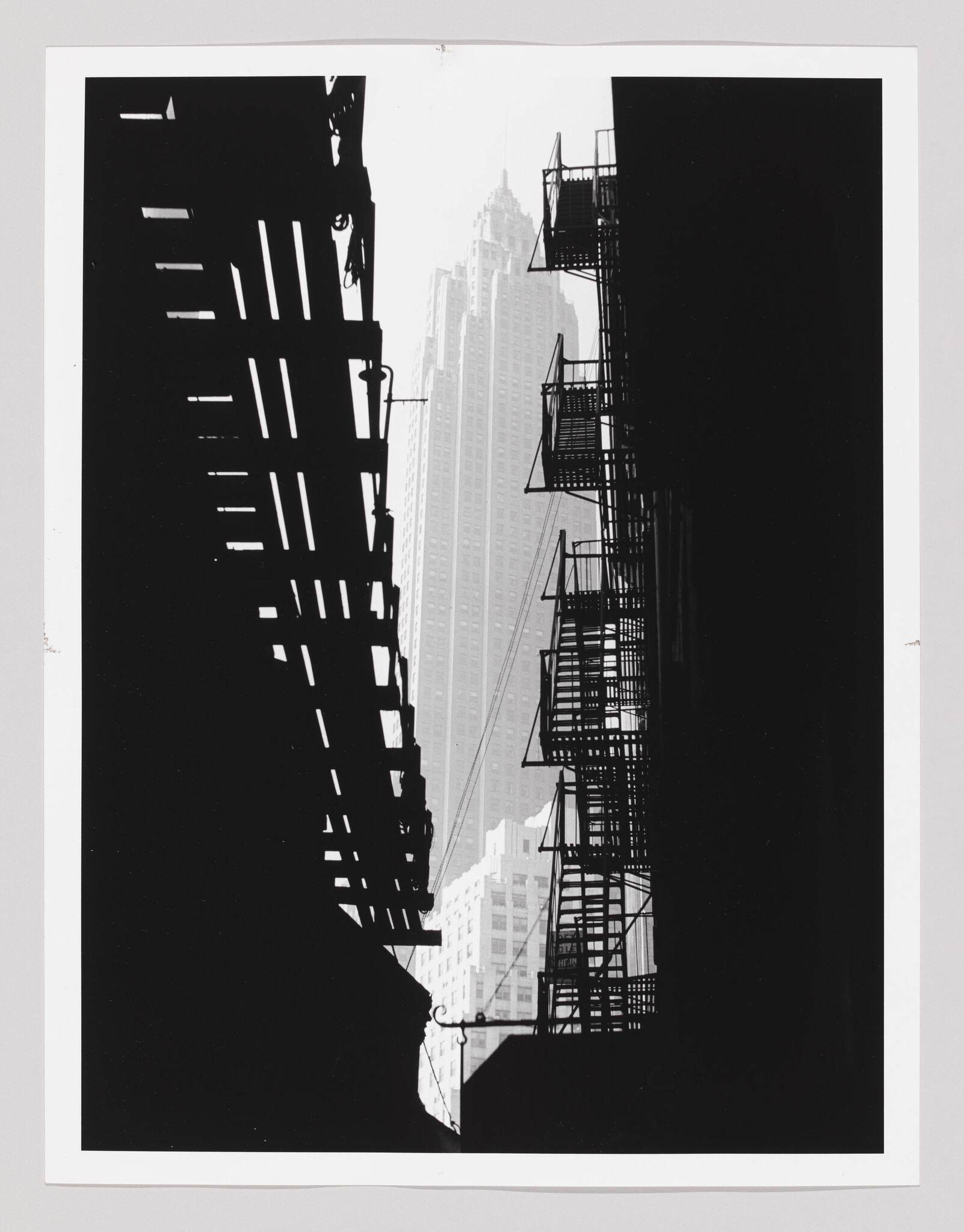 Silhouetted fire escapes and elevated train tracks frame a distant skyscraper in the city.