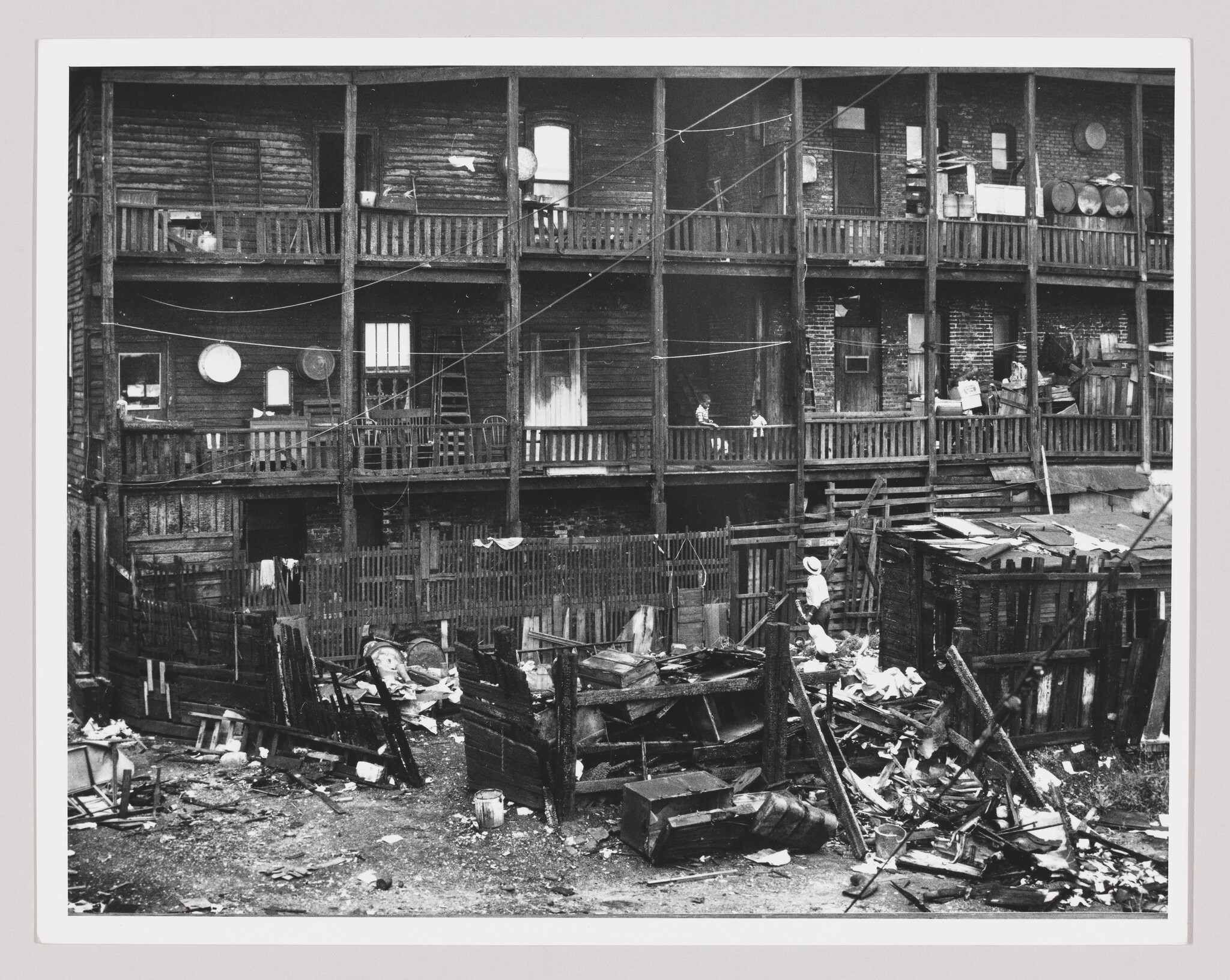 Three-story wooden tenement with cluttered backyard full of debris and a person walking near rubble.