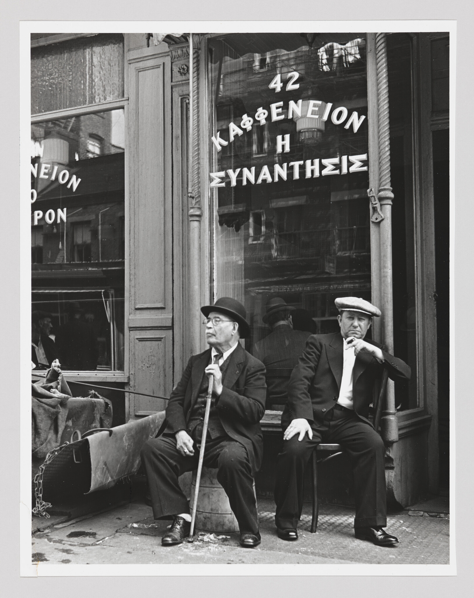 Two older men sit outside a café, one holding a cane and the other smoking a cigarette.