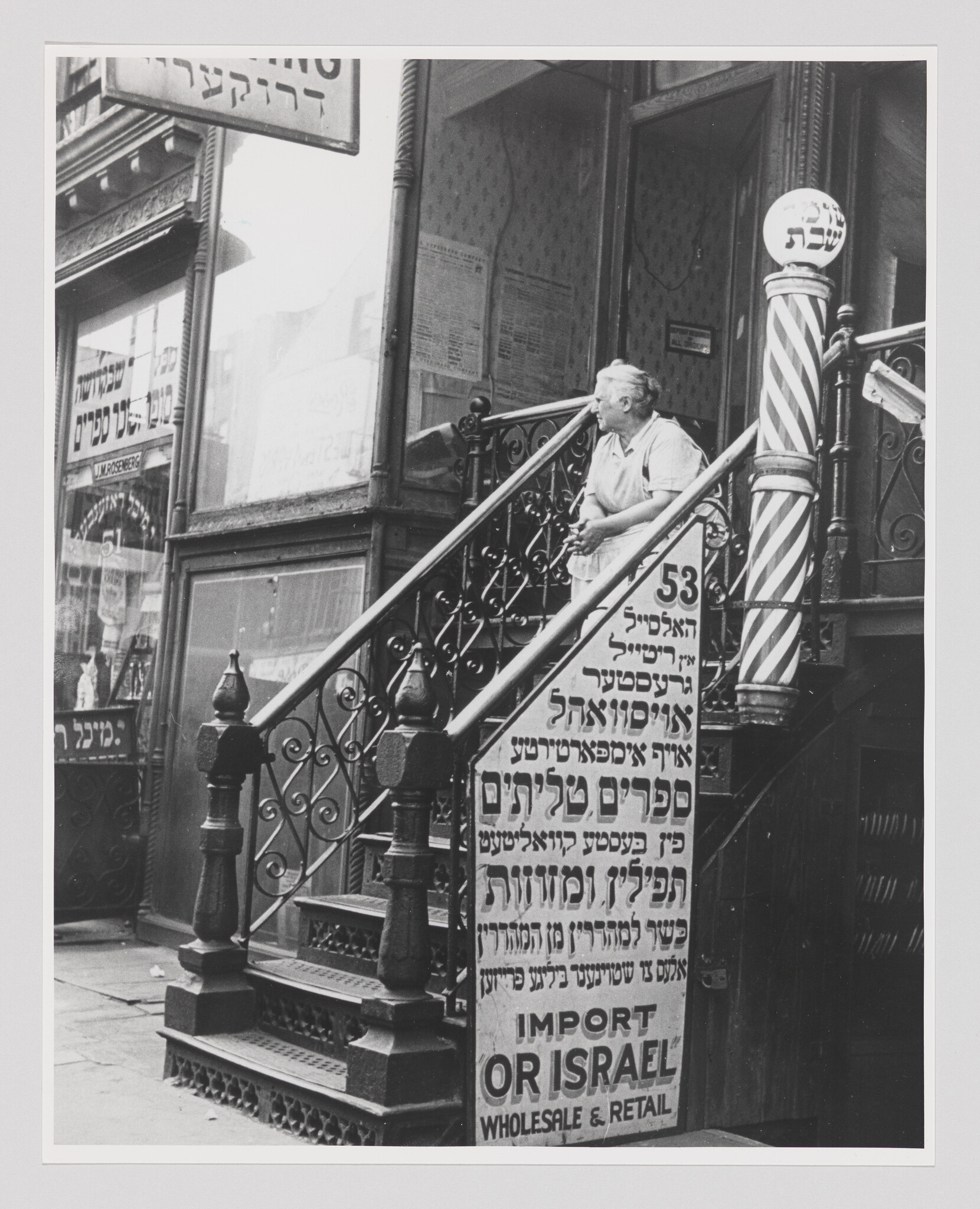 An elderly woman stands on a storefront stoop beside a sign reading "Import or Israel."