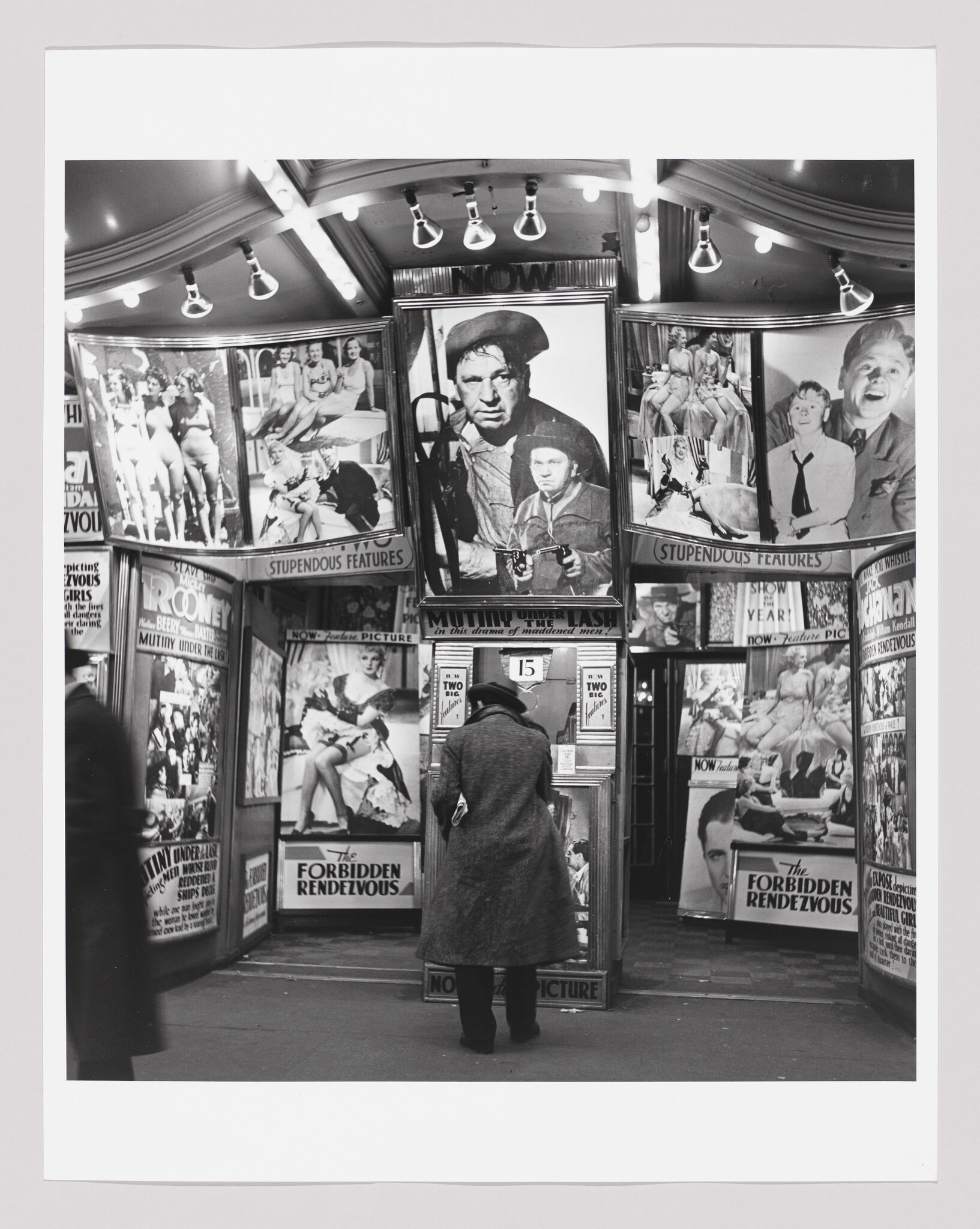 An individual in a coat leans at a ticket booth surrounded by large movie posters and lights.