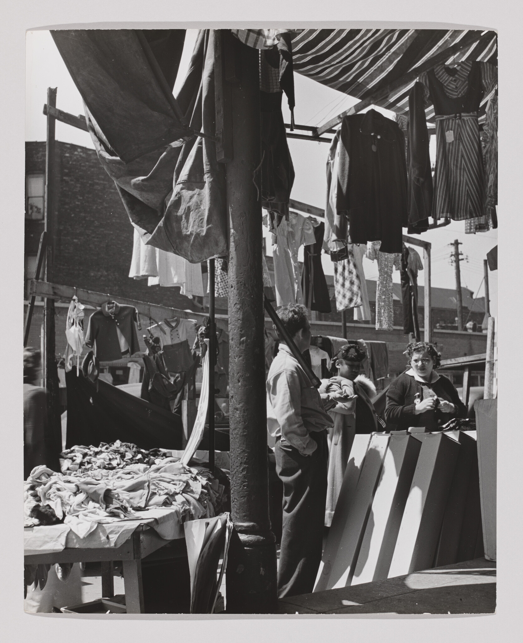 Three people talk at a busy outdoor clothing stall with garments hanging overhead.