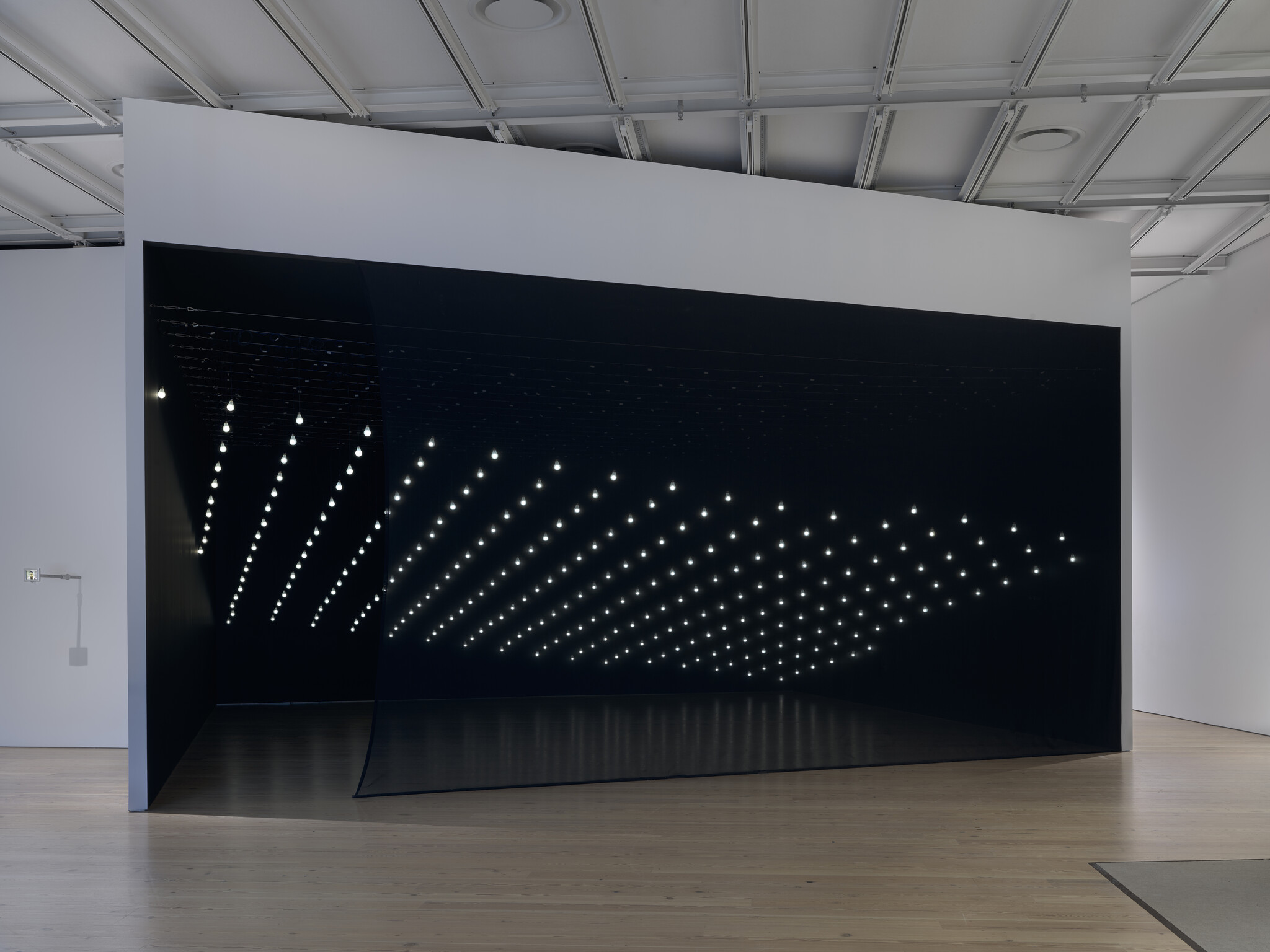 Large dark room installation with rows of small white lights hanging from the ceiling in a geometric pattern.