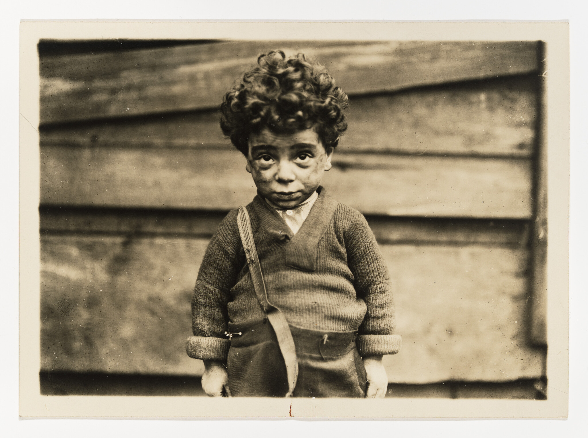 Vintage sepia-toned photograph of a doll with human-like features, wearing a knitted sweater and trousers, standing in front of a wooden slat background. The doll has curly hair and a somewhat somber expression.