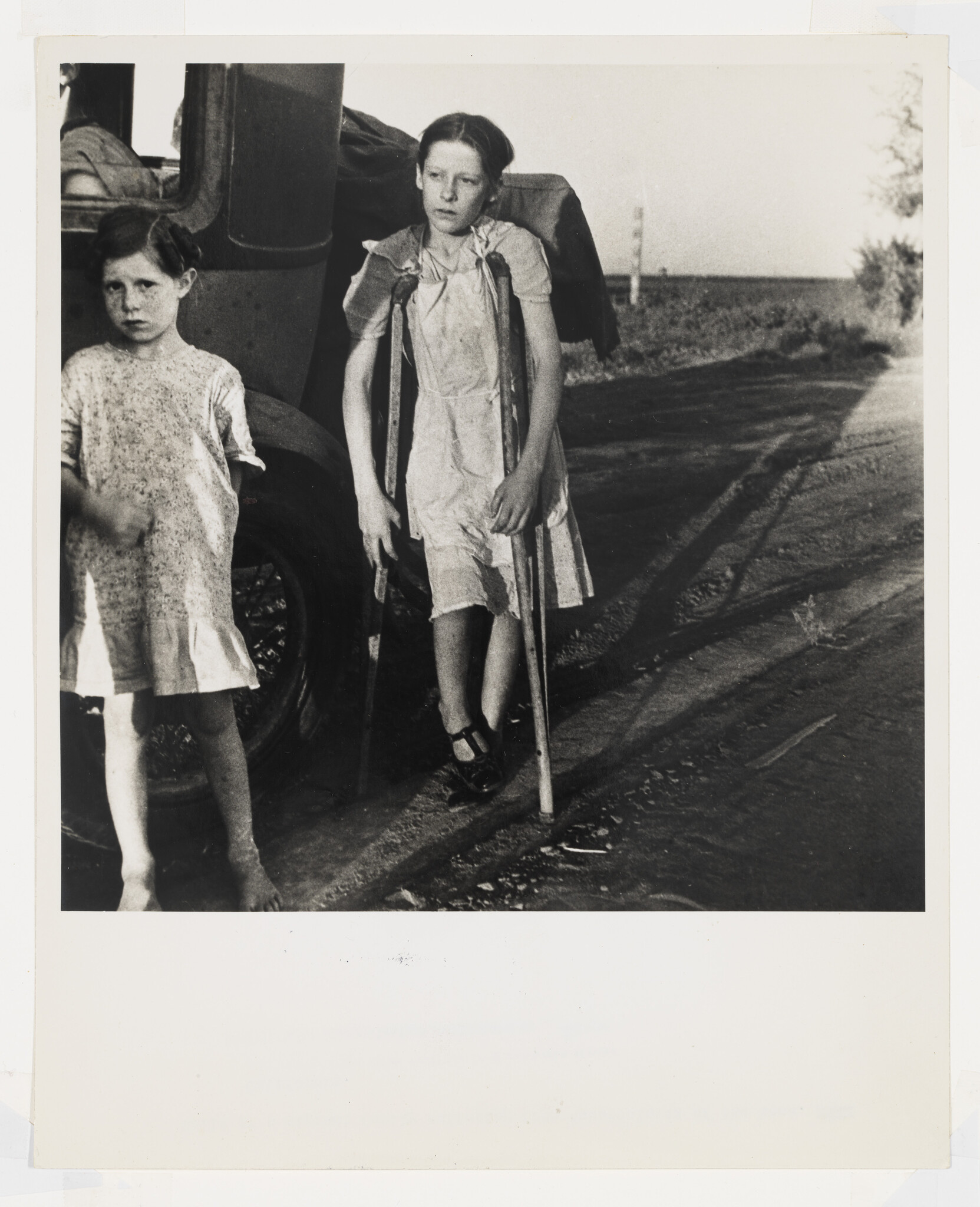 Two young girls stand beside a car on a rural road, one using crutches to walk.