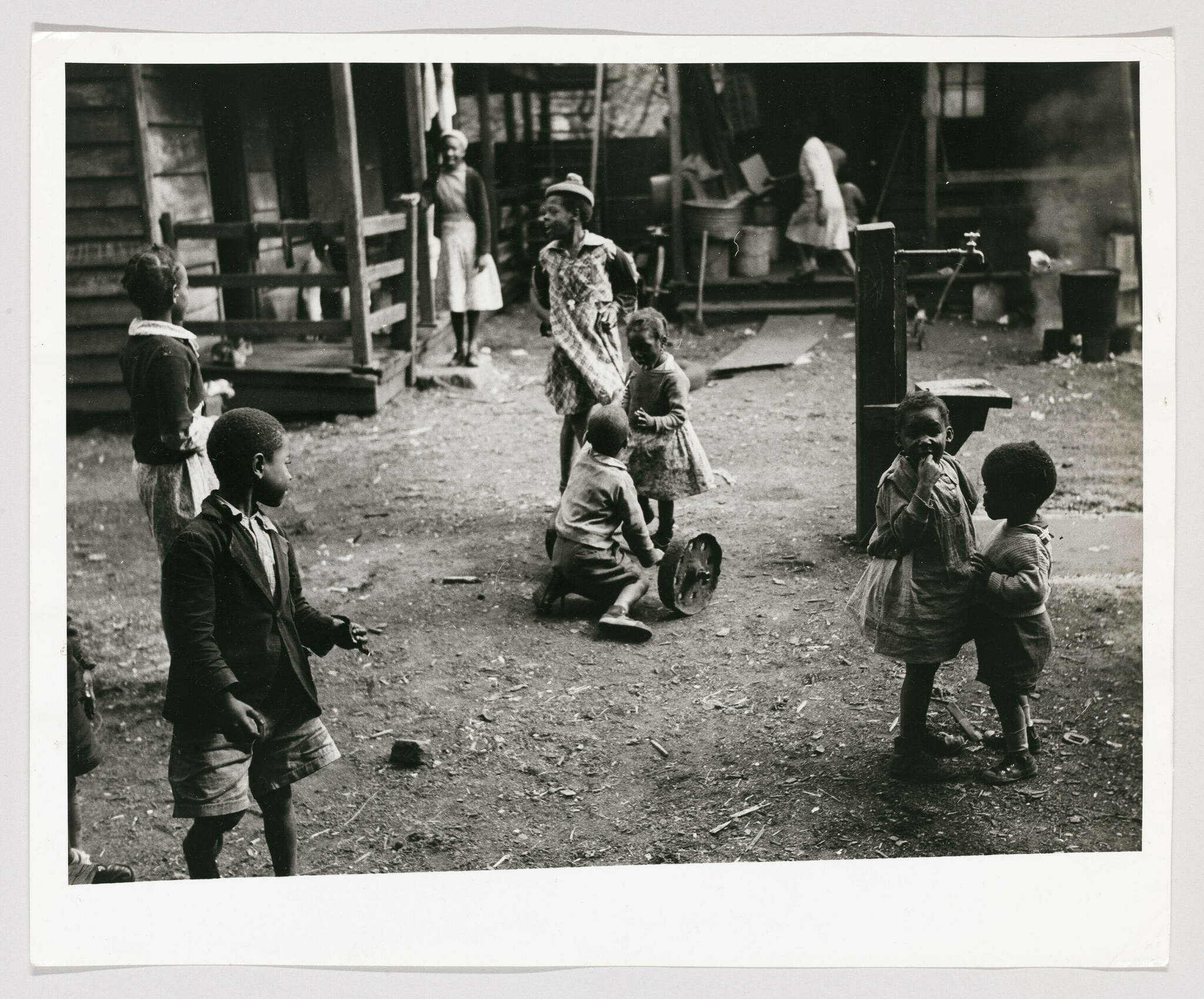 A black and white photograph depicting a group of African American children playing in a dirt yard in front of wooden houses. Some children are standing and looking around, while others are sitting on the ground. An adult woman is visible in the background, standing on a porch, and another is working near a pile of buckets. The scene suggests a communal living area, possibly from an earlier time period, with a rustic and informal setting.