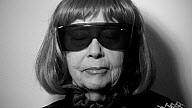 An older person wearing large dark sunglasses and a short bob wig looks straight ahead.