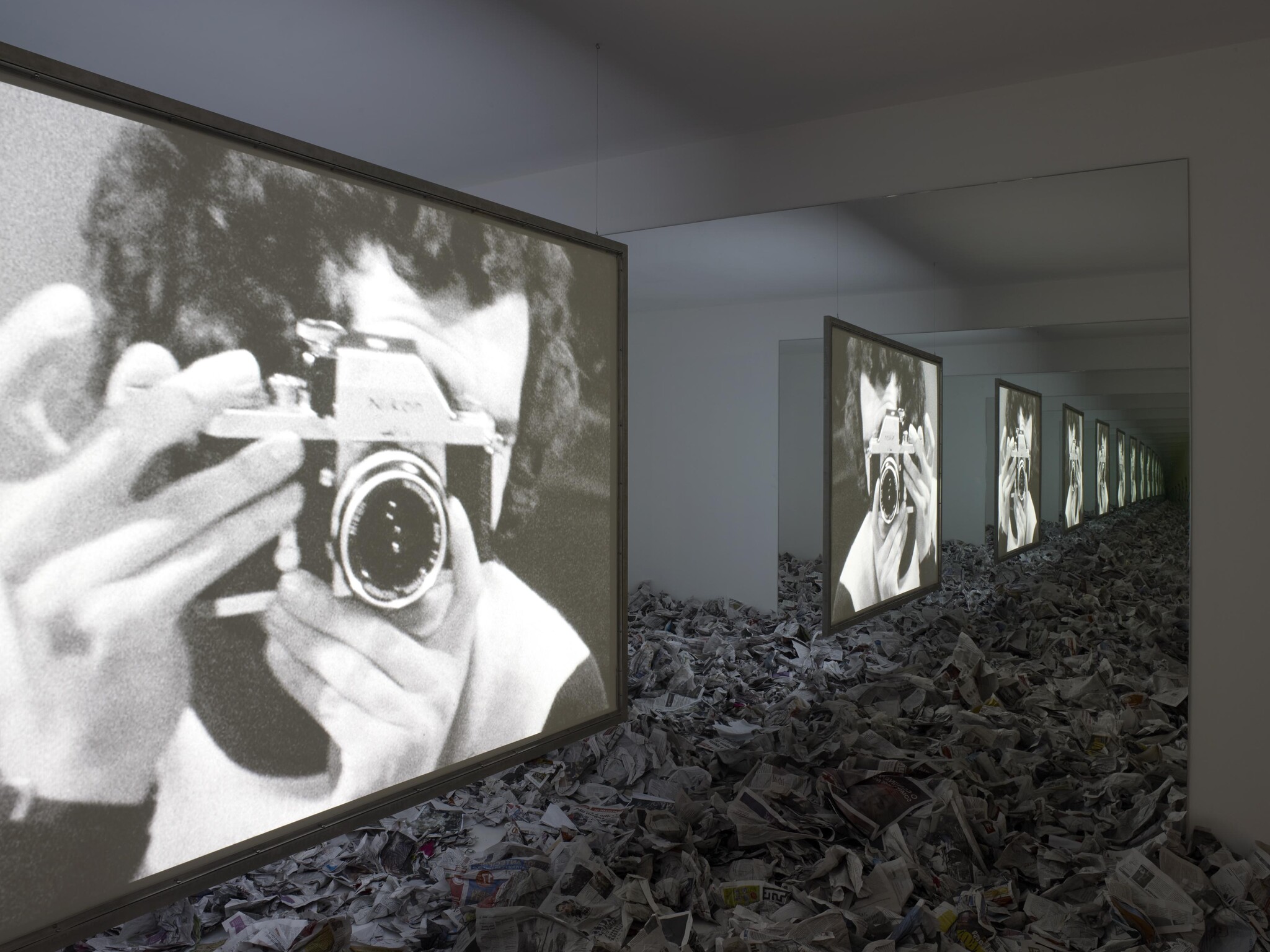 Large screens show a person holding a camera while a room floor is piled with newspapers.