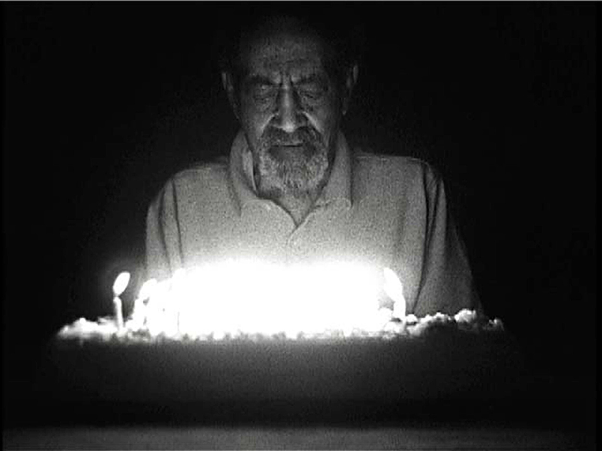 An elderly bearded man looks down at a brightly lit birthday cake with candles.
