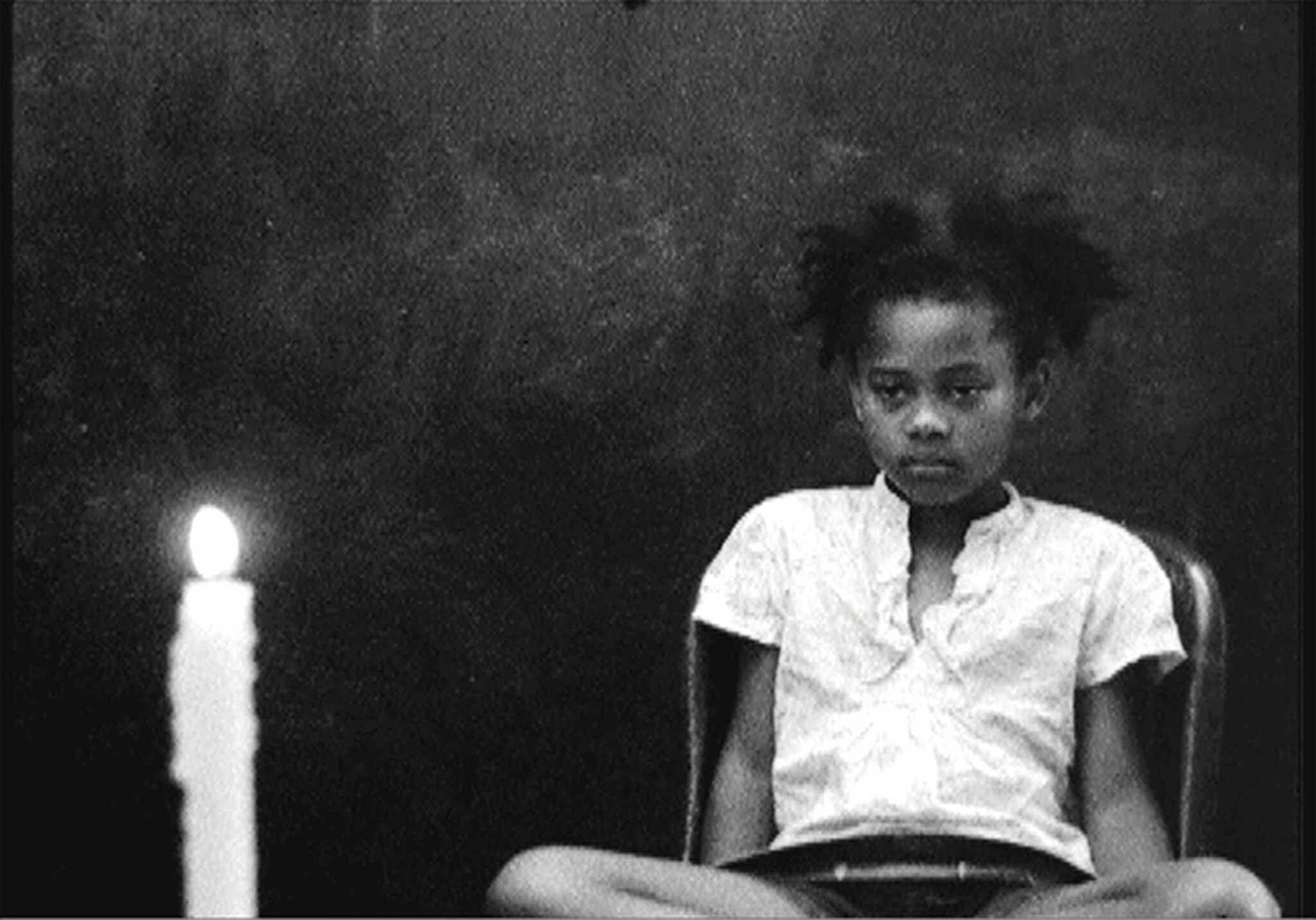 A young girl sits solemnly on a chair while a lit candle burns beside her.