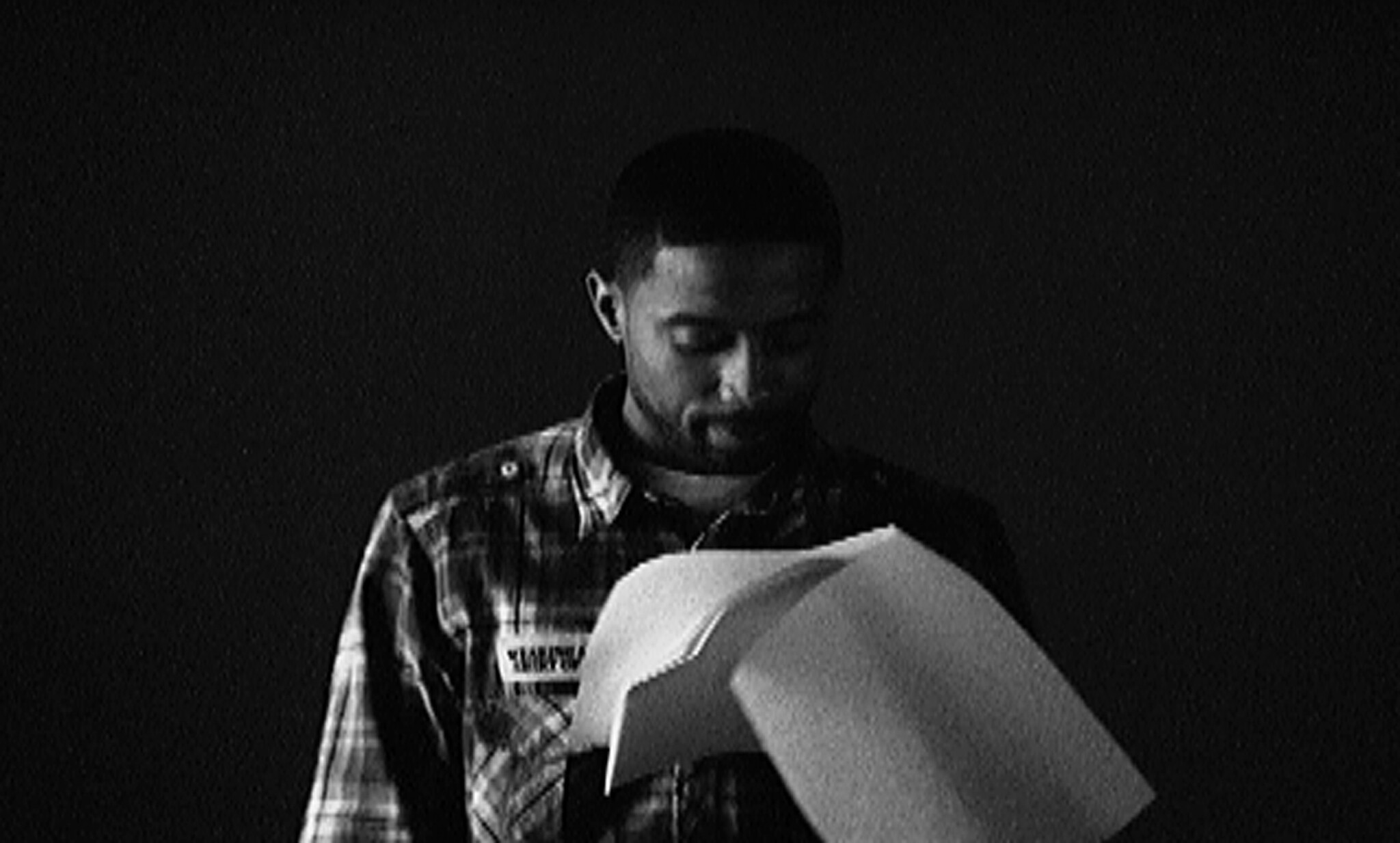 A man in a plaid shirt looks down while reading loose papers against a dark background.