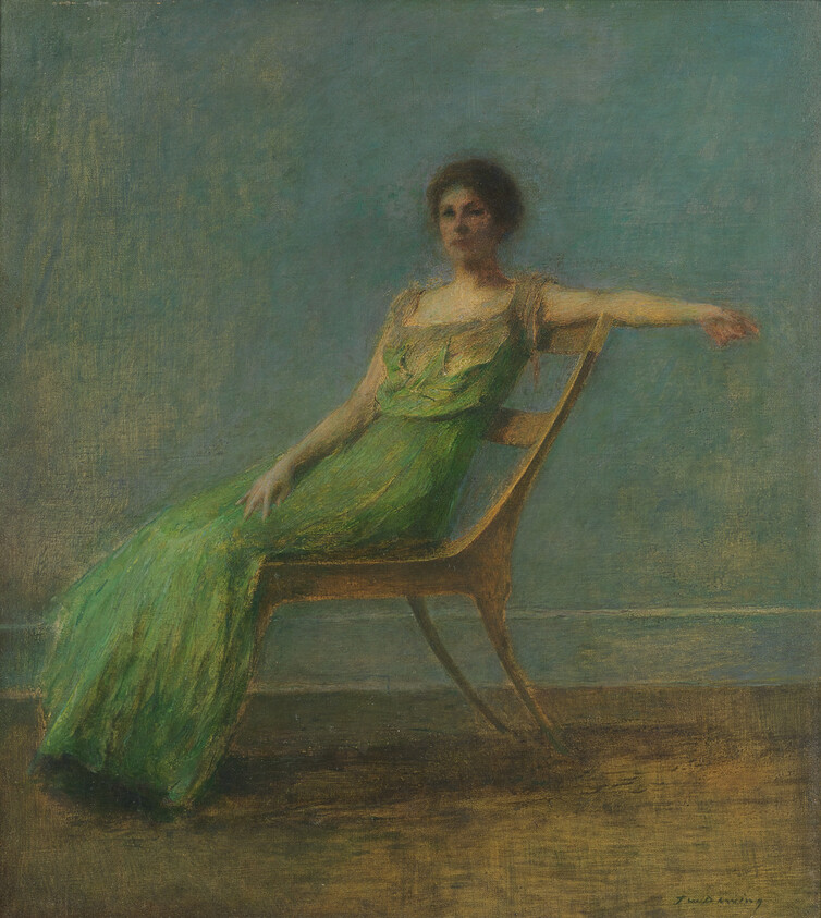 A woman in a long green dress reclines casually on a wooden chair against a muted wall.