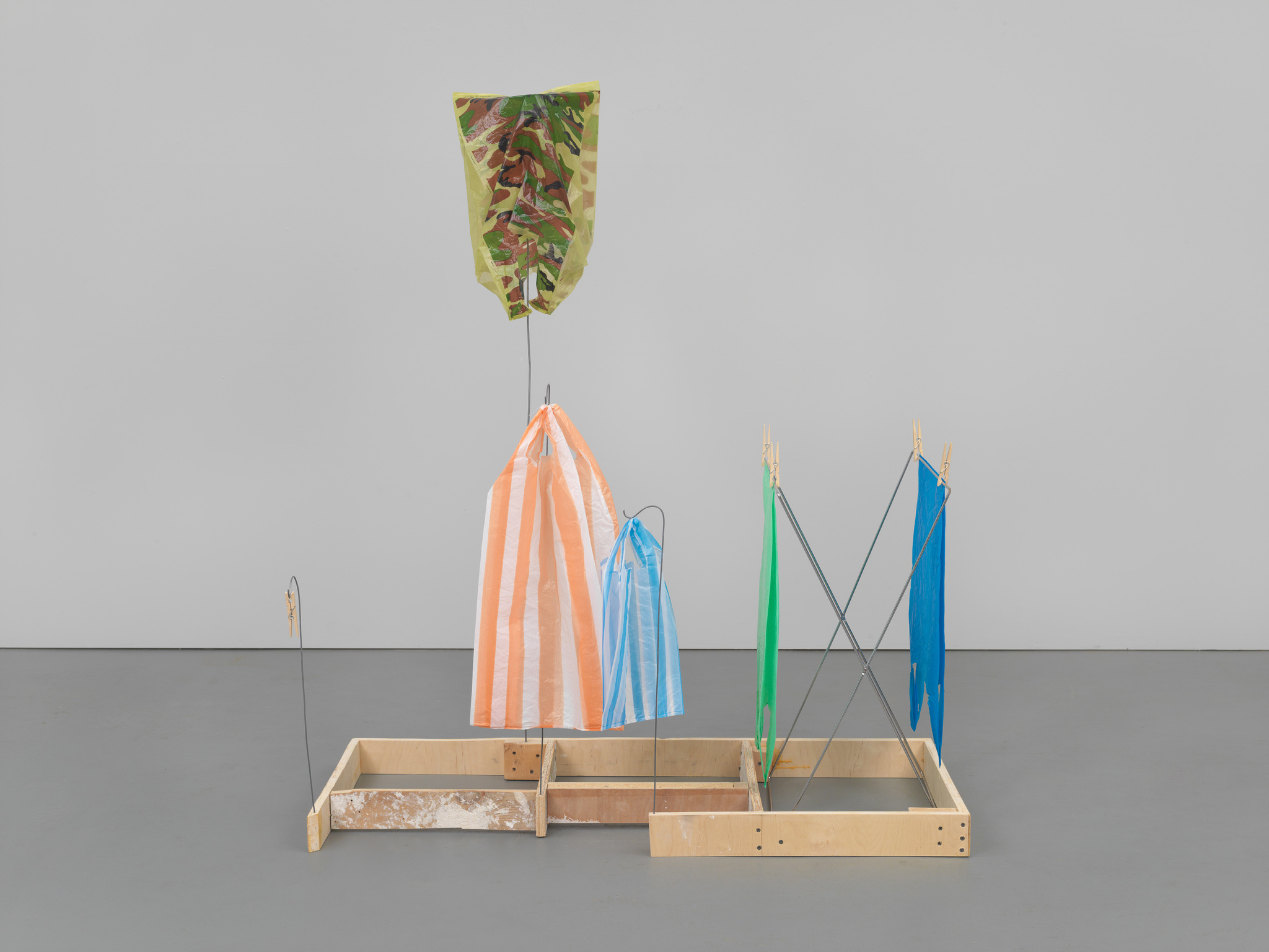 A contemporary art installation featuring colorful garments suspended from metal frames, with a camouflage kite hanging above, all set against a plain gray floor and white wall background.