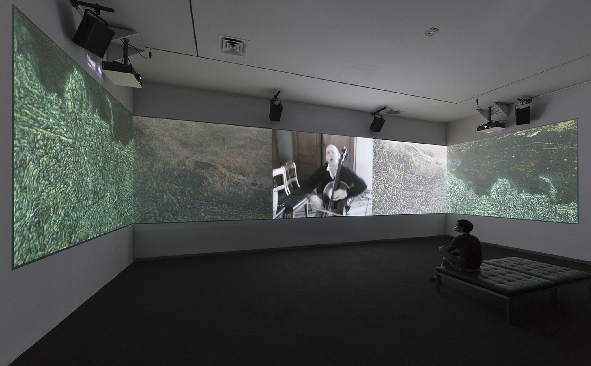 A person sits on a bench watching a large multi-screen projection of a cellist performing.