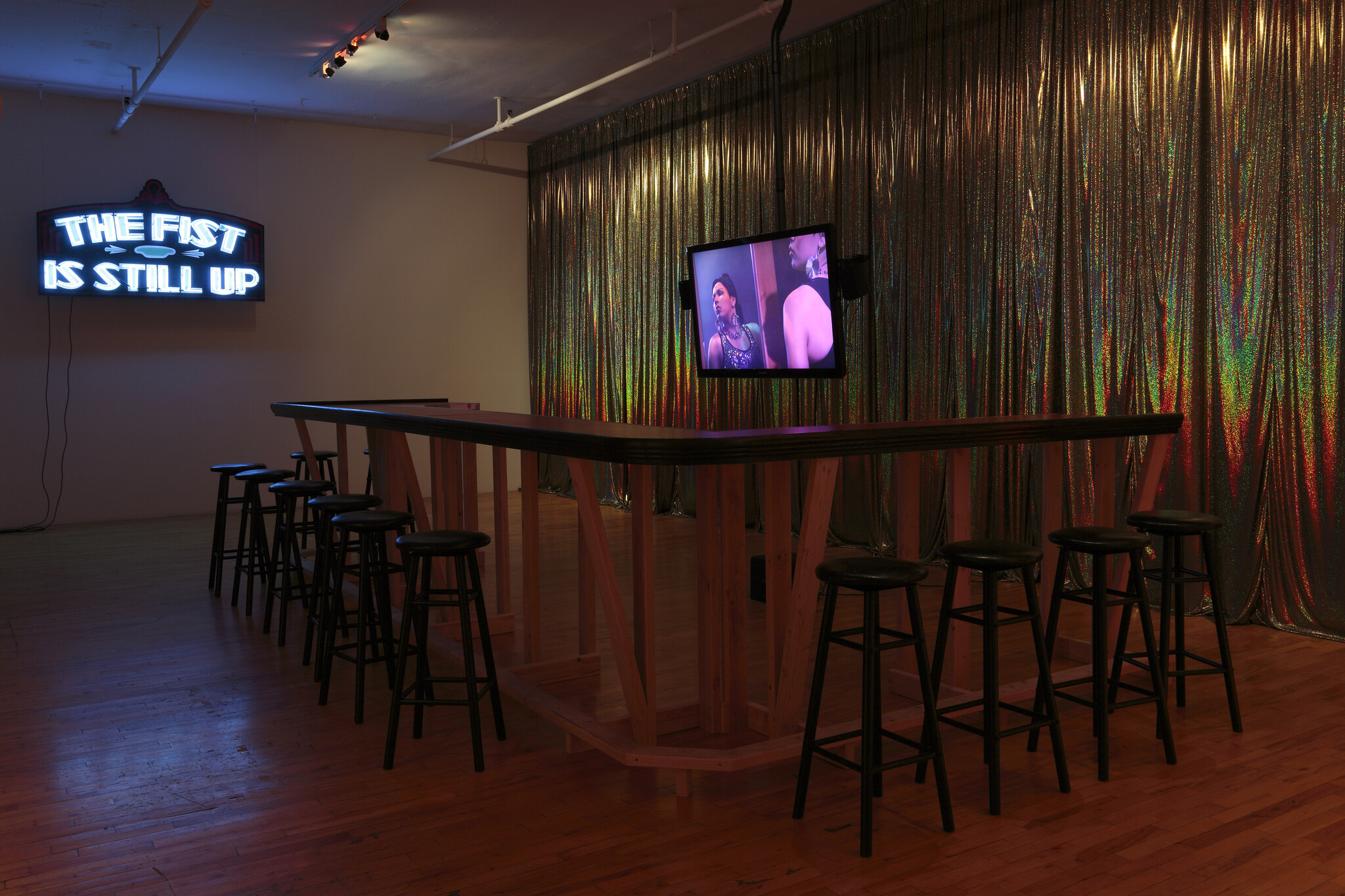 A long wooden bar with stools sits under a neon sign reading "The fist is still up."