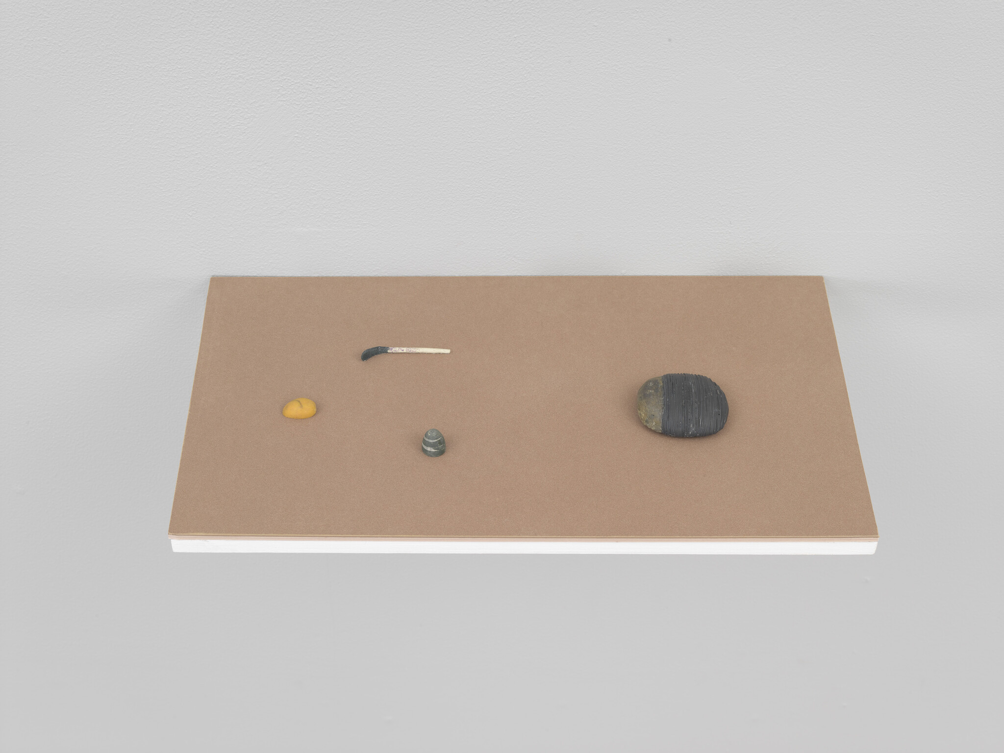 A minimalist art installation featuring a brown rectangular surface with five small objects arranged asymmetrically: a yellow blob, a bent nail, a small greenish stone, a tiny screw, and a larger black and yellow striped stone, against a white wall.