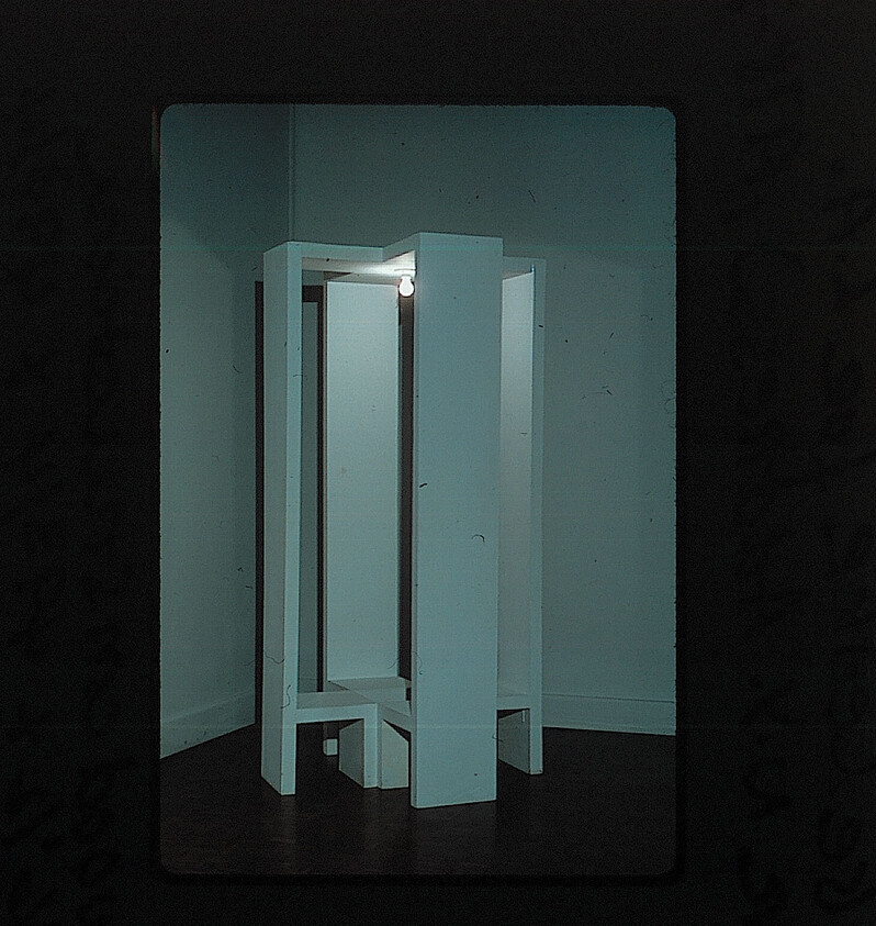 Tall, white, geometric structure with open sides stands in a corner, lit by a single exposed bulb.