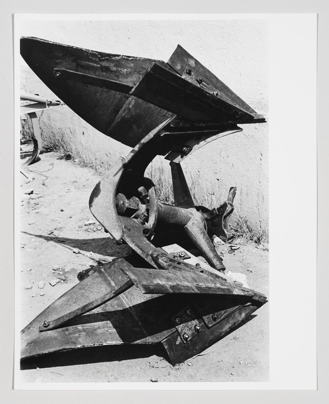 A black and white photograph of a large, abstract metal sculpture resembling a flower with multiple layers of petals, displayed outdoors with a textured wall in the background.