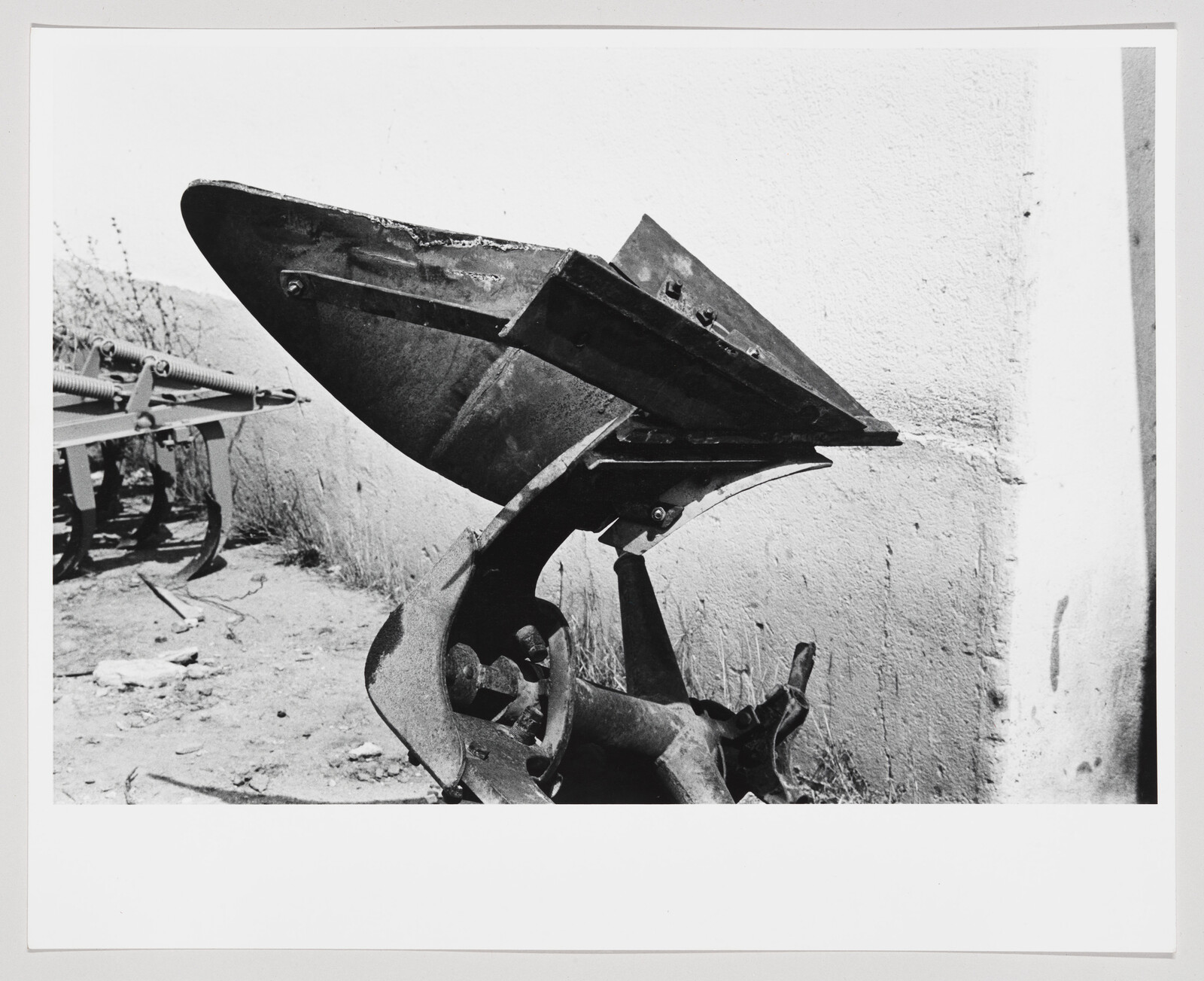 A black and white photograph showing a close-up of a large, metallic, rusted agricultural plow blade with its mechanism, set against a backdrop of a white wall and some sparse vegetation.