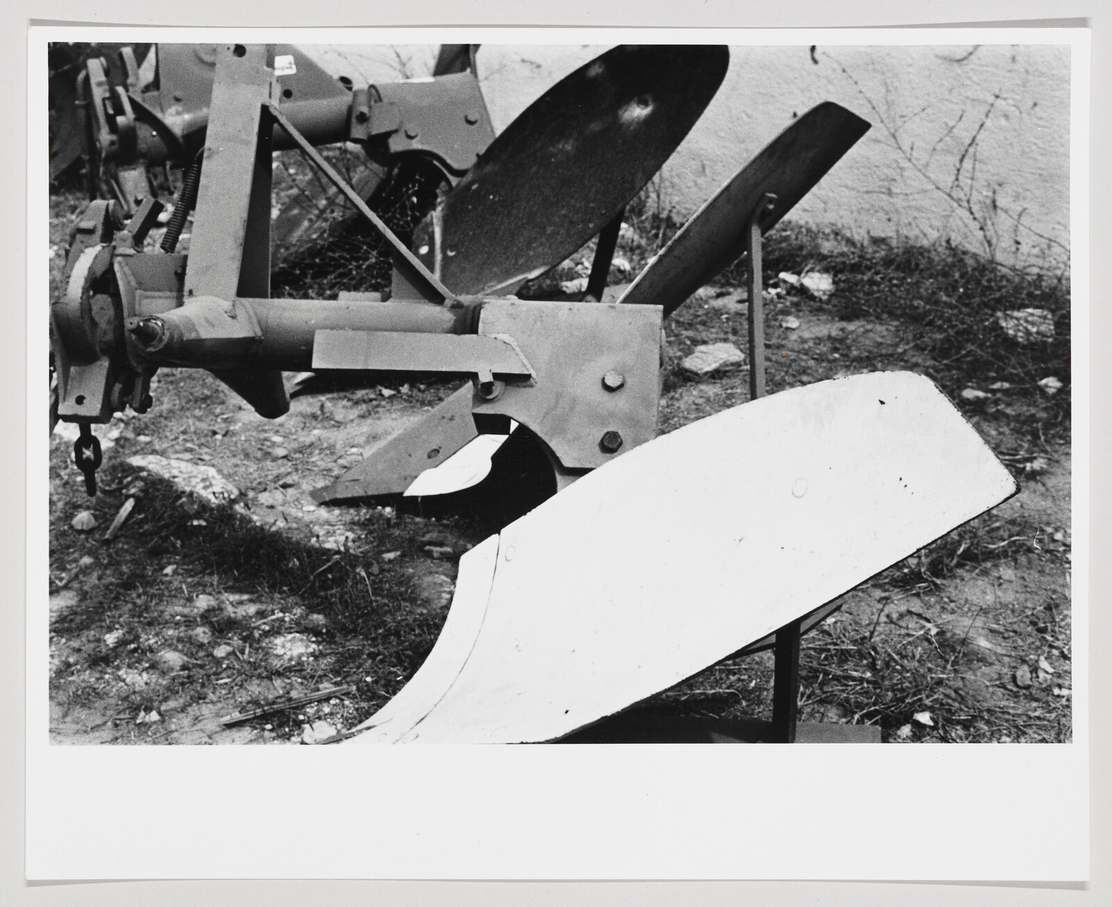 Black and white photograph of a plow with multiple blades and mechanical parts, positioned on a patch of ground with sparse grass and a wall in the background.
