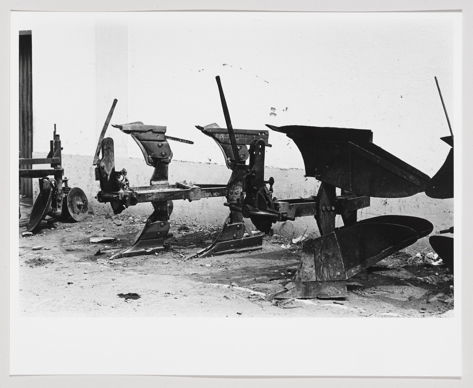 Black and white photograph showing a series of old, metal printing presses with large flywheels and levers, arranged in a row in a room with a dirty floor and white walls. The equipment appears aged and not in use.