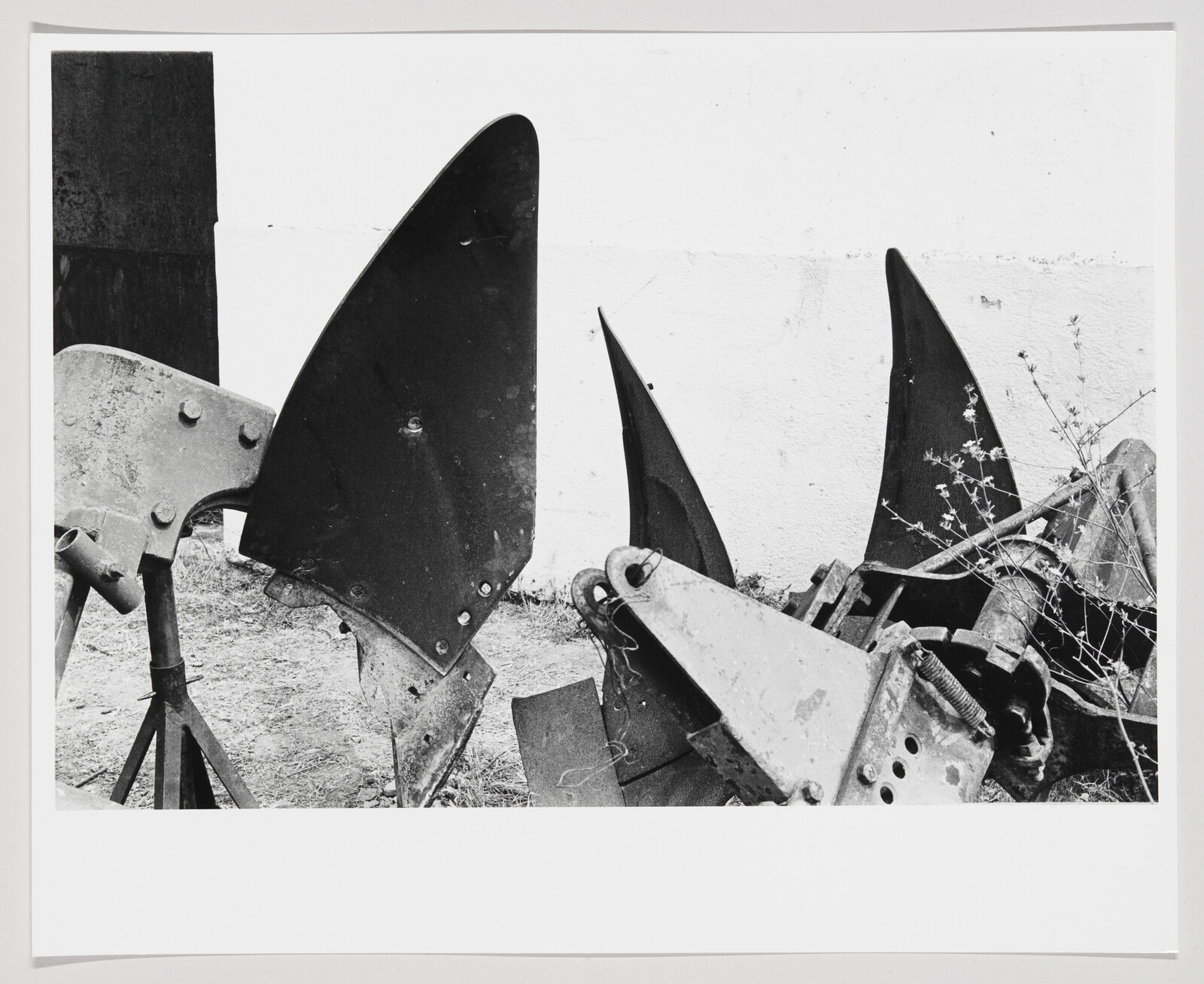 A black and white photograph showing a collection of discarded metal plow blades and agricultural equipment piled against a wall.
