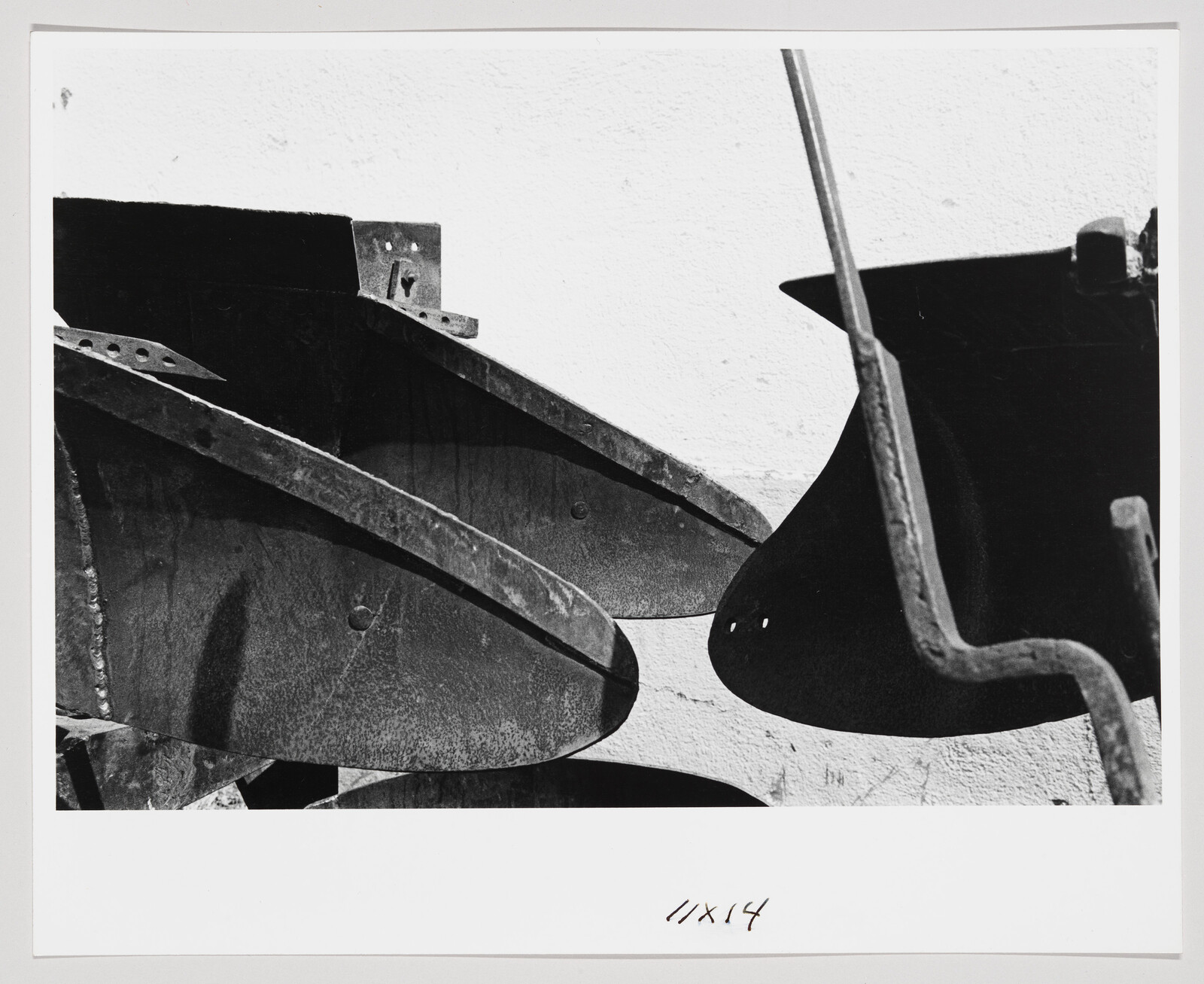 A black and white photograph featuring abstract shapes and shadows of what appears to be metal sculptures or parts, with a stark contrast between the dark figures and the light background. The composition is dynamic, with angular and curved elements interacting. There is handwriting at the bottom right corner of the image that reads "11x14".