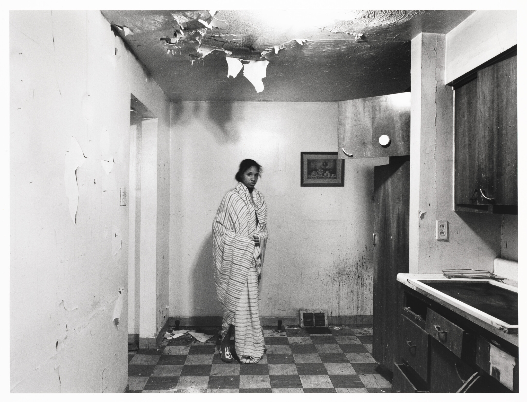 Woman wrapped in a blanket stands in a rundown kitchen with peeling ceiling and floor tiles.