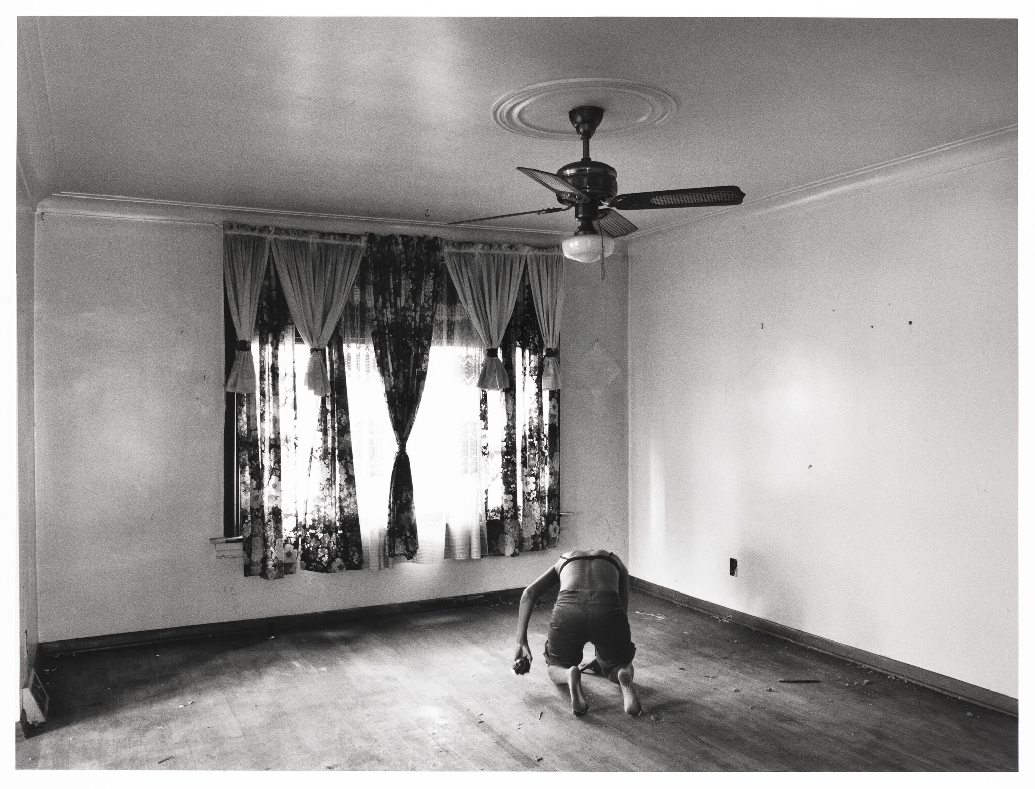 A person is kneeling and examining the empty room's bare floor near floral curtains.