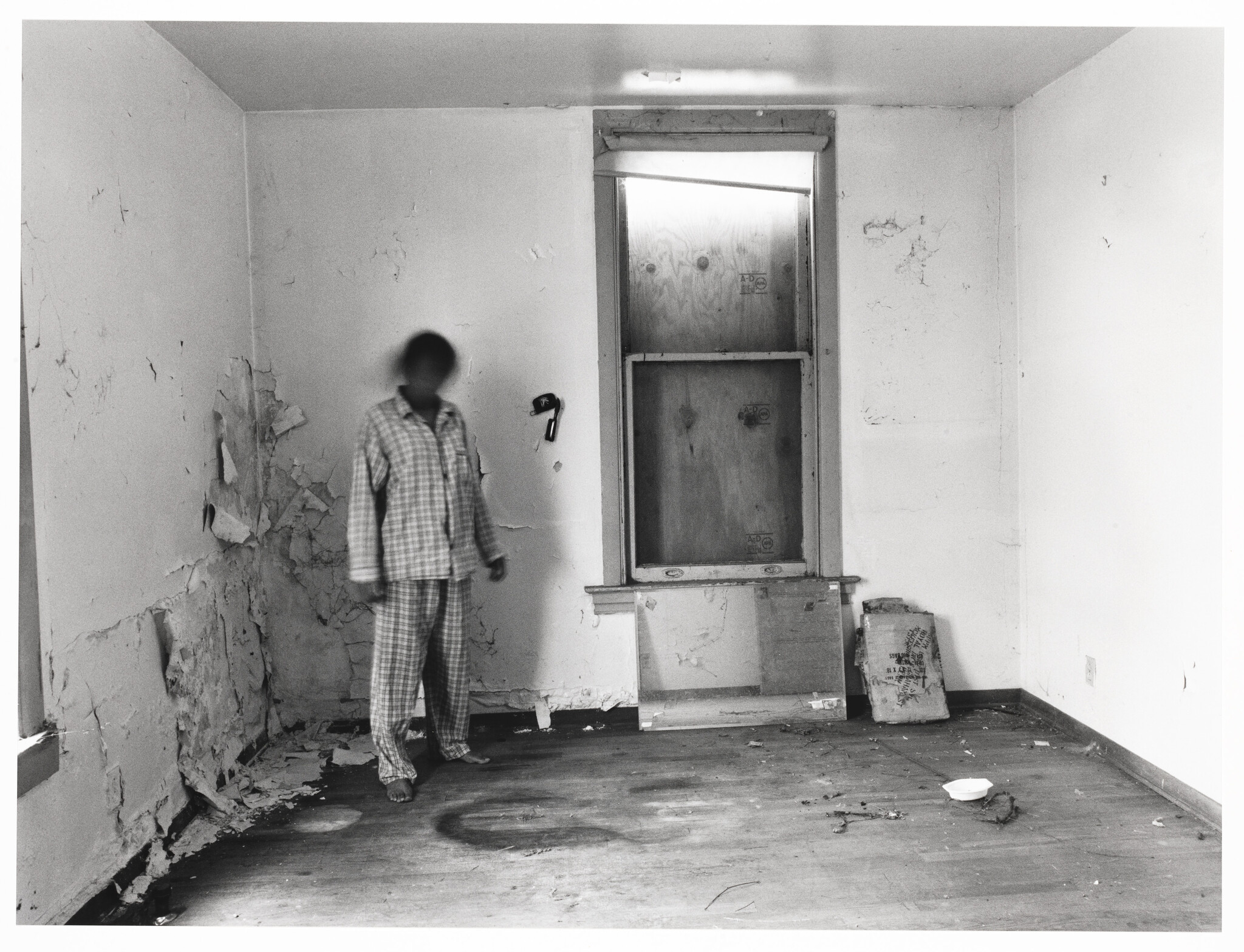 A barefoot person in plaid pajamas stands in a dilapidated, empty room with a boarded window.
