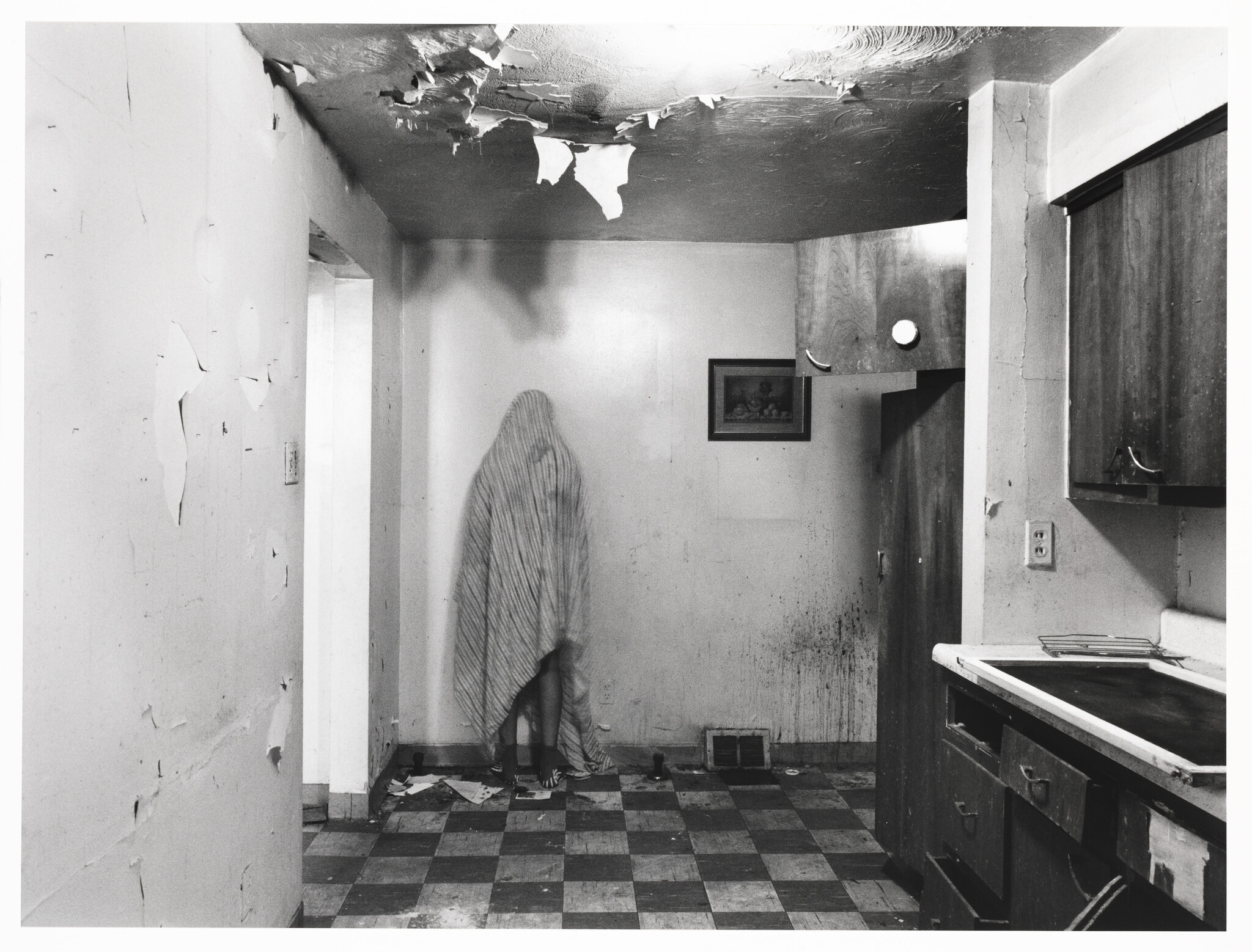 A black and white photograph depicting a dilapidated kitchen with peeling paint and a damaged ceiling. In the corner stands a person obscured by a patterned blanket, creating a ghostly appearance. The room has a checkered floor, and there's a small picture frame on the wall. The overall atmosphere is eerie and abandoned.