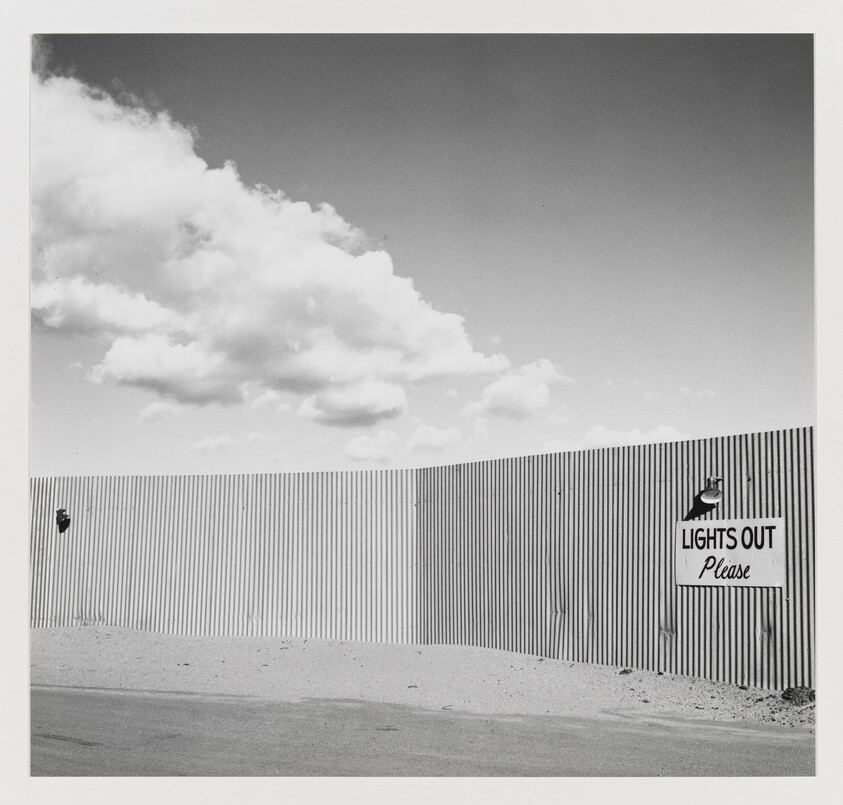 A corrugated metal fence with a sign reading "Lights out Please" under a cloudy sky.