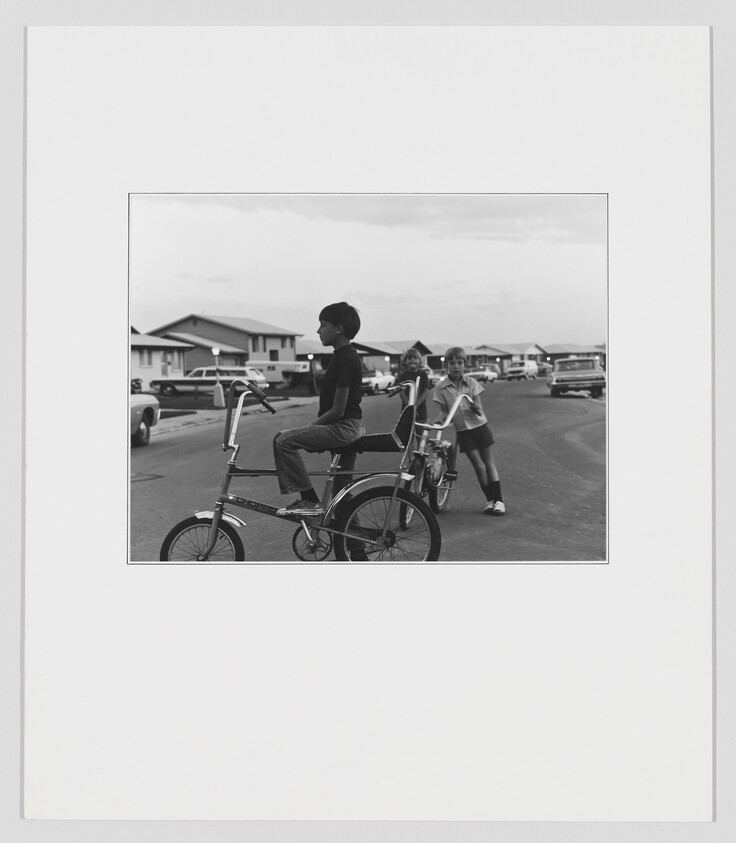 A black and white photograph capturing a moment from the past where a young boy sits atop a tall, custom bicycle, looking into the distance. Behind him, a girl is walking with a bicycle, both are in a residential area with houses and parked cars in the background. The image is mounted on a white border, giving it a framed appearance.