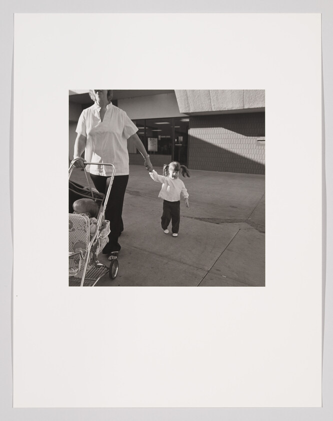 Adult holds a toddler's hand while pushing a stroller with a baby outside a building.
