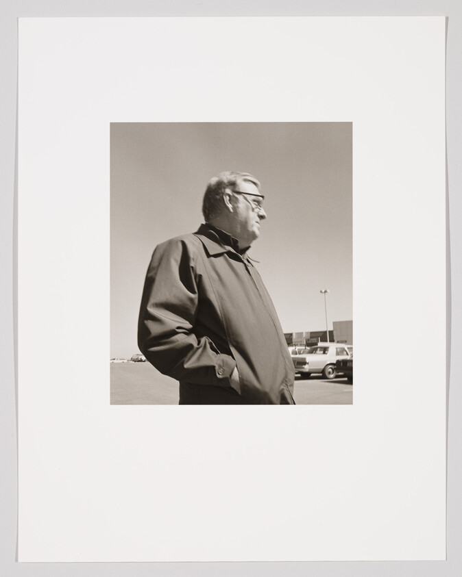 An older man stands in a parking lot with his hands in his pockets, looking to the right.