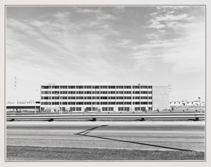 A long four-story office building sits behind a highway guardrail under a wide cloudy sky.