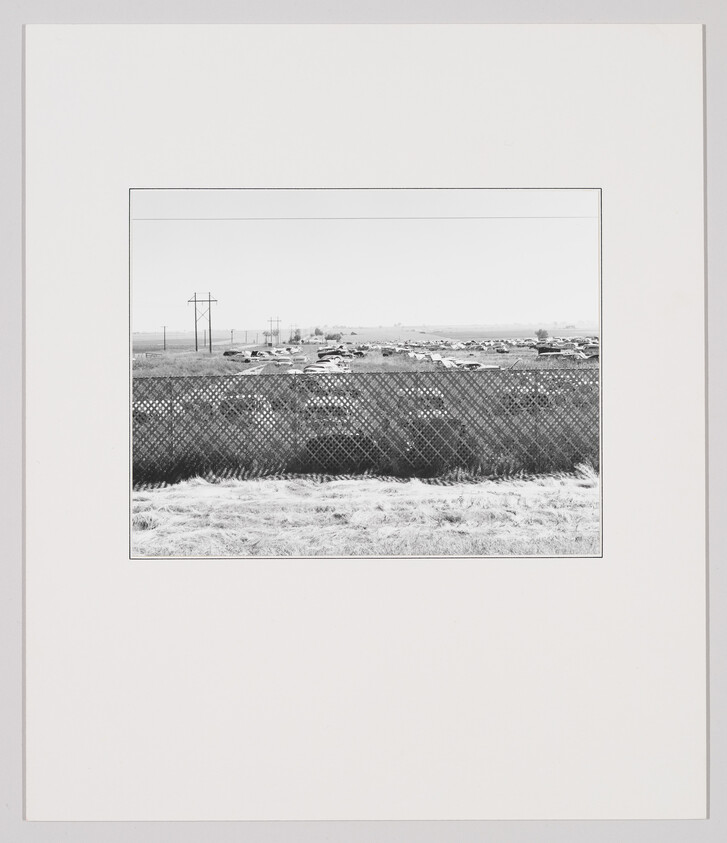 A chain-link fence in front of a grassy field and rows of parked cars under power lines.