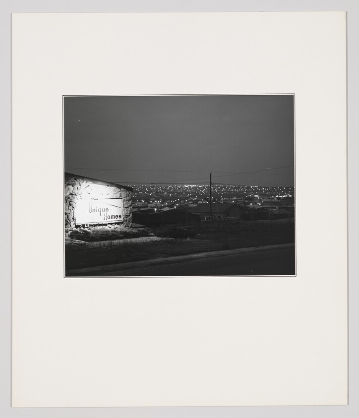 A lit stone sign reading "Unique Homes" overlooks a sprawling, illuminated suburban neighborhood at night.