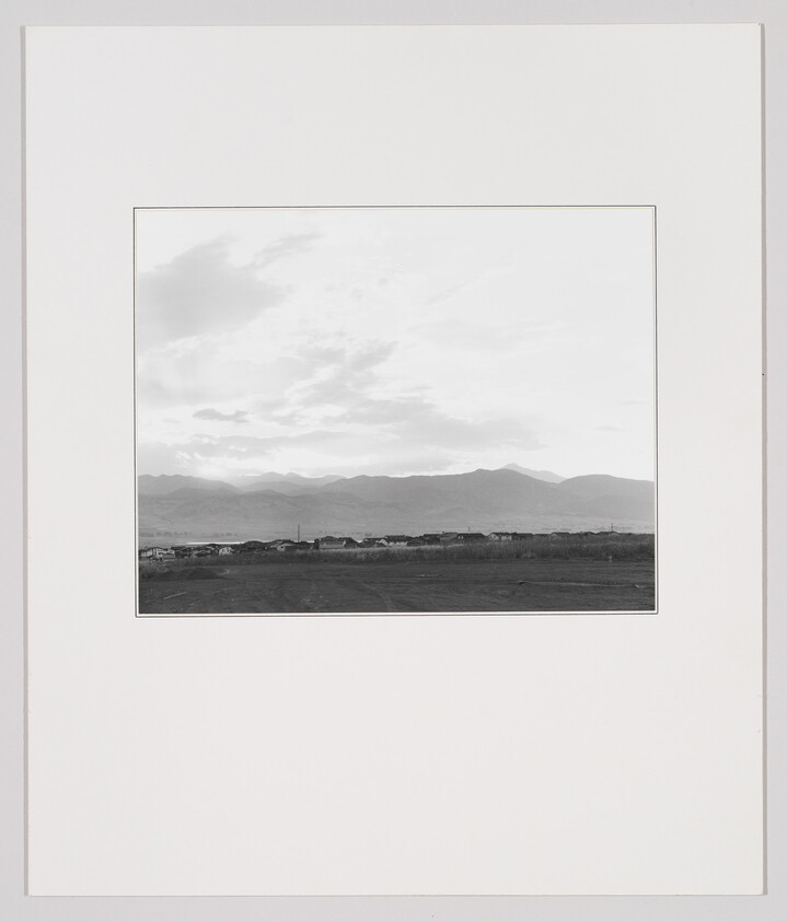 Distant mountain range above a low row of houses with open fields and cloudy sky.