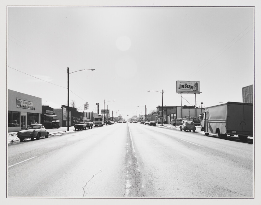 A wide, empty main street lined with parked cars and a Jim Beam billboard overhead.