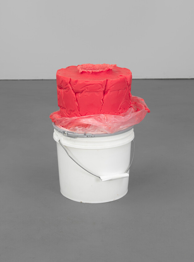 A white plastic bucket holds a large lump of red wax wrapped in clear plastic.