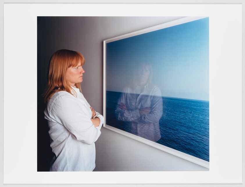 A woman stands with folded arms, studying a framed photograph of the ocean reflecting her image.